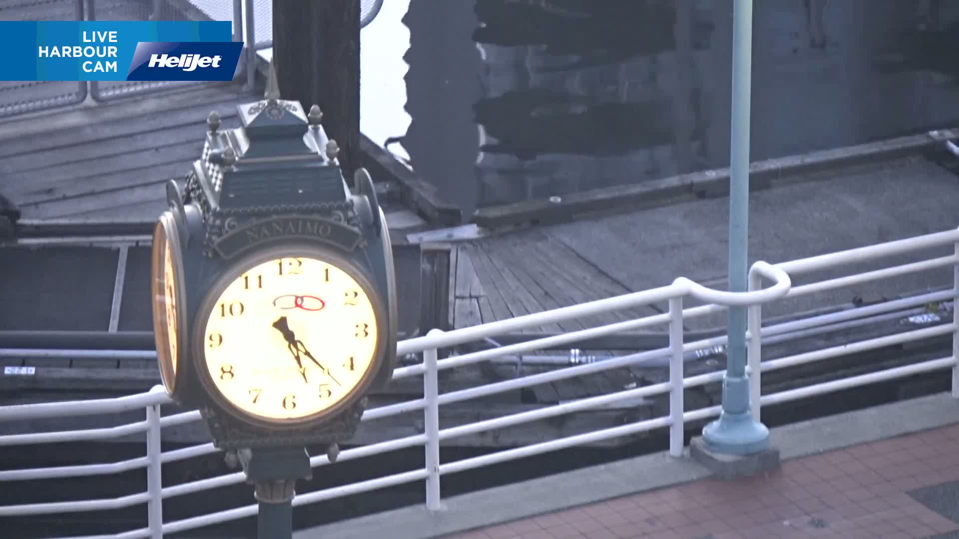A lit clock on a post stands beside a white railing overlooking a dark body of water on an overcast day.