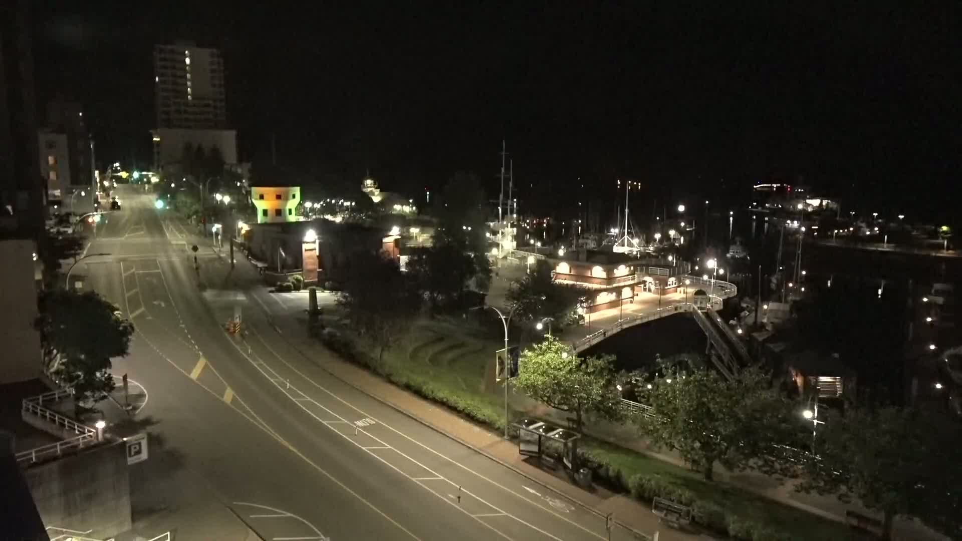 A nighttime, high-angle view shows a mostly empty road curving past a marina with docked boats and a park, all lit by streetlights.