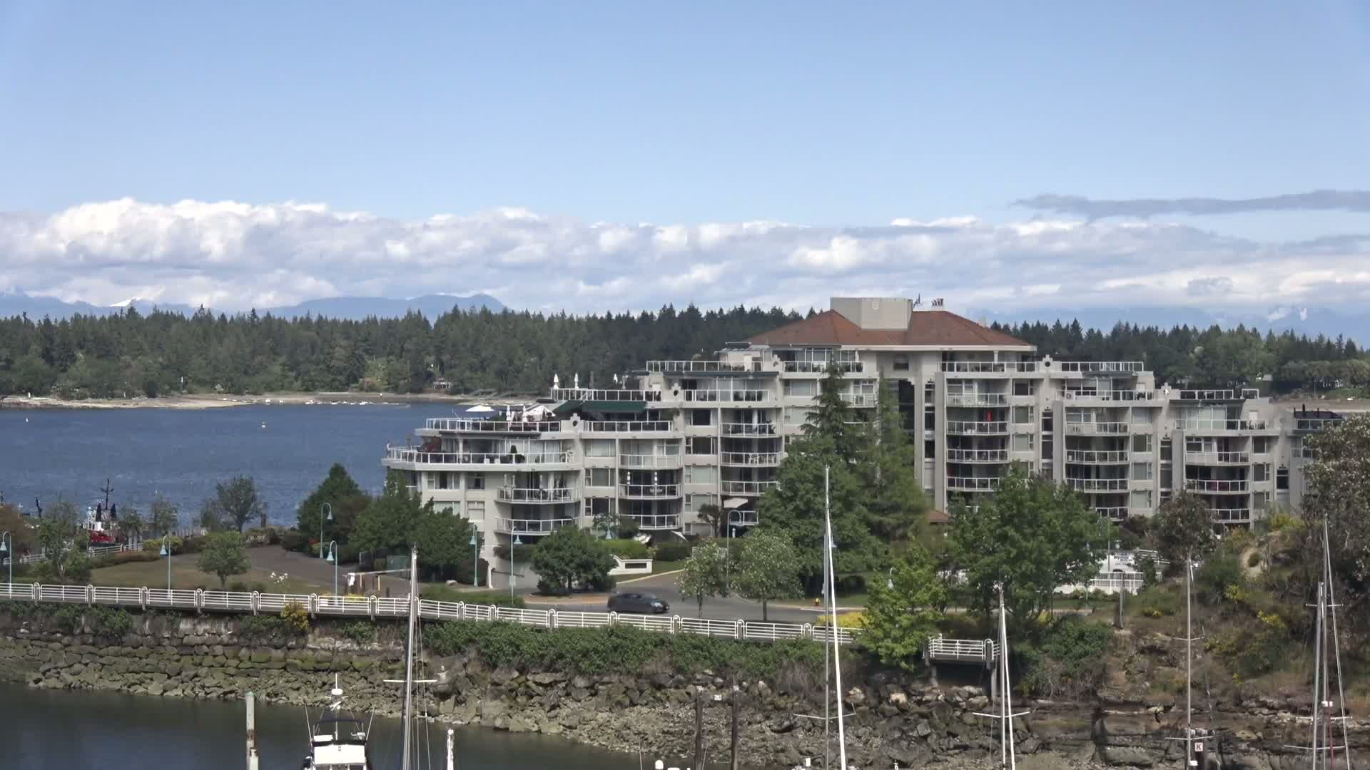 A large, multi-story building sits on a waterfront, partially obscured by trees and sailboat masts, under a partly cloudy sky.