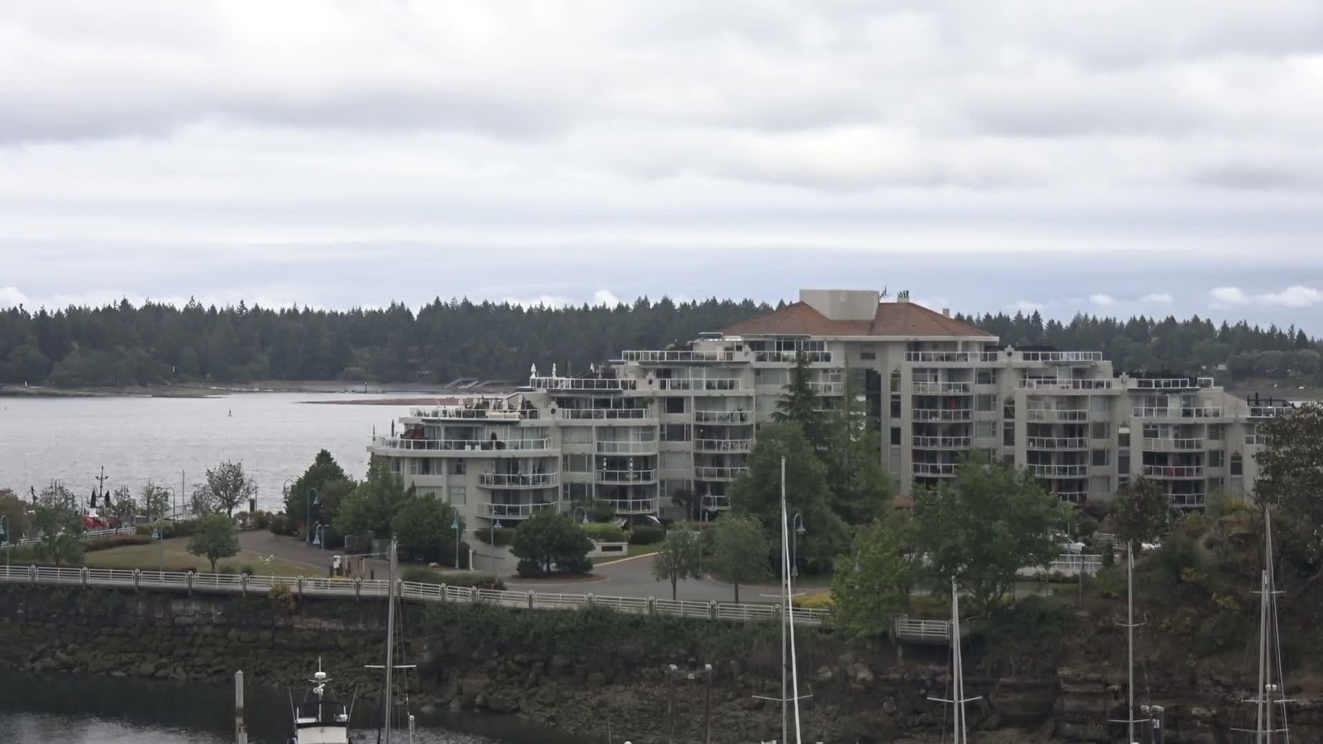 A large multi-story apartment building sits on a waterfront, surrounded by trees and a calm body of water under a cloudy sky.
