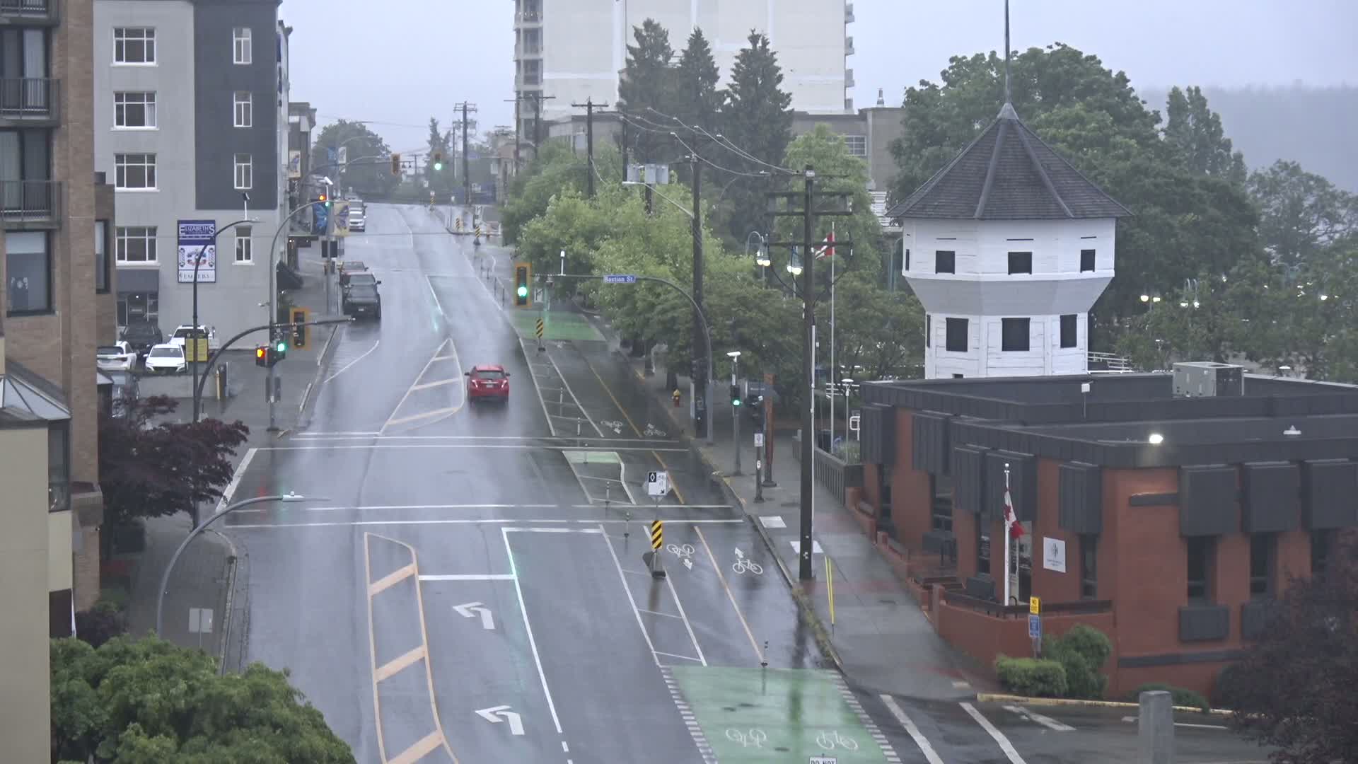 A wet city street scene on a rainy day shows a multi-lane road with bike lanes, several cars, and buildings including a multi-story apartment building and a unique octagonal tower attached to a brick building.