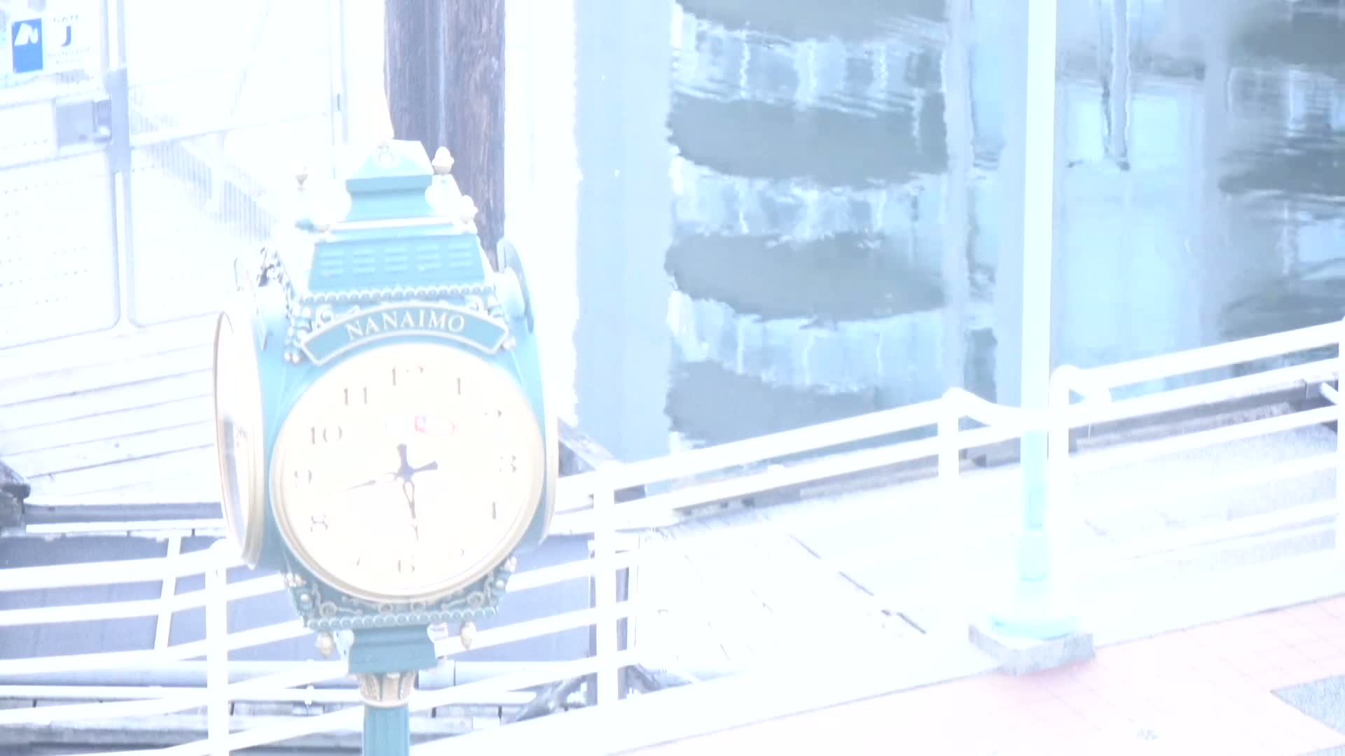 A teal street clock with a gold face shows the time on an overcast day next to a white railing overlooking calm water.
