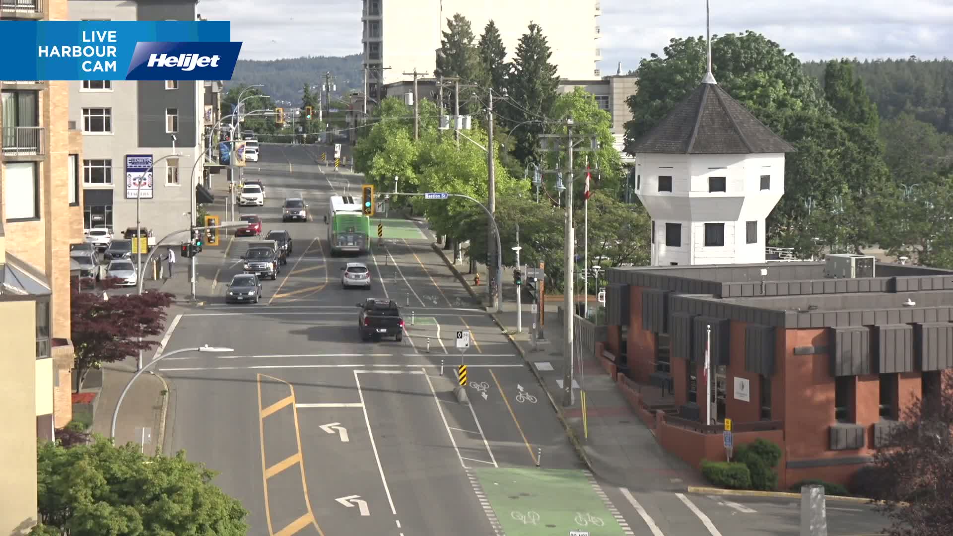 A sunny day shows a city street intersection with several cars, a bus, and a white octagonal tower attached to a brick building, all surrounded by lush green trees.