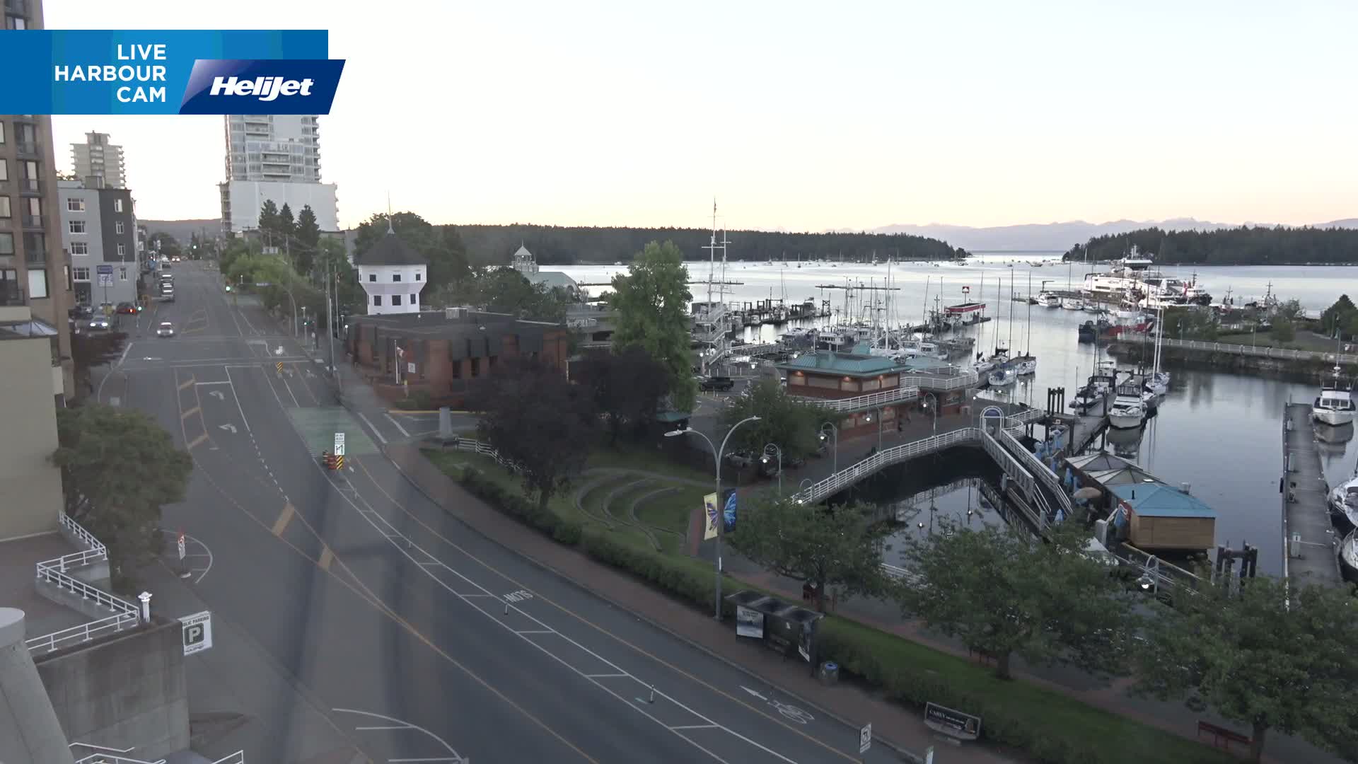 A mostly empty harbor with numerous boats and a quiet roadway next to a green space, under a partly cloudy sky.