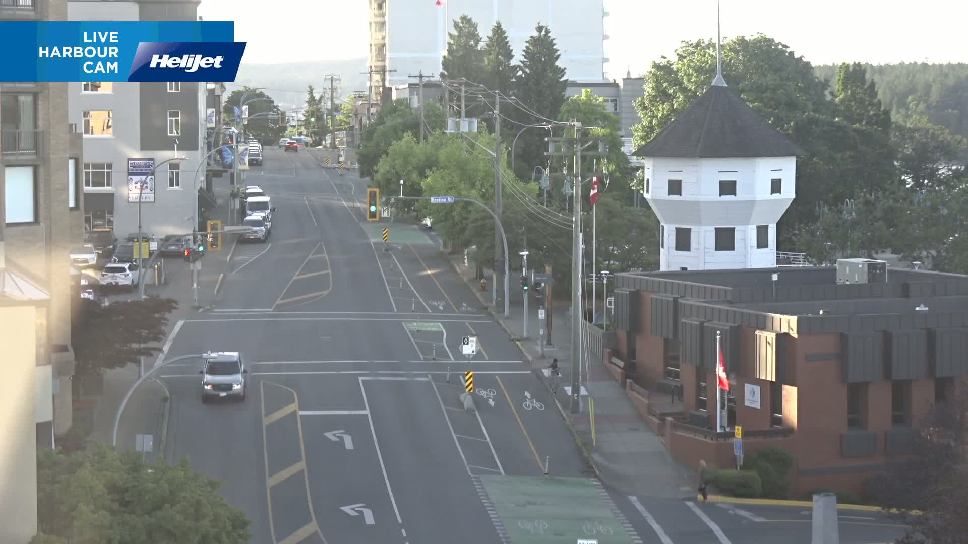 A mostly empty street scene on a sunny day features a white octagonal tower next to a brick building, with cars sparsely scattered along the road and bike lanes visible.