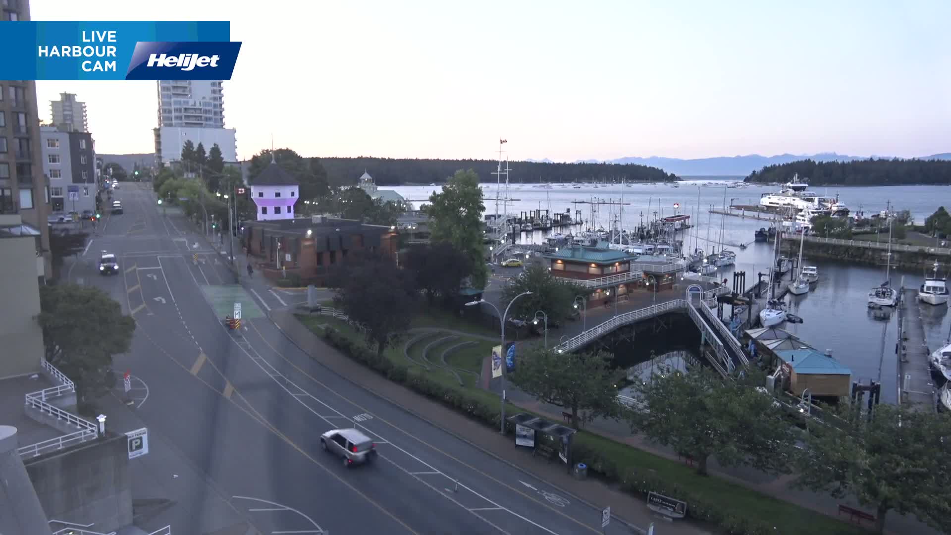 A harbor with numerous boats docked, a road with a few cars, and buildings under a twilight sky.