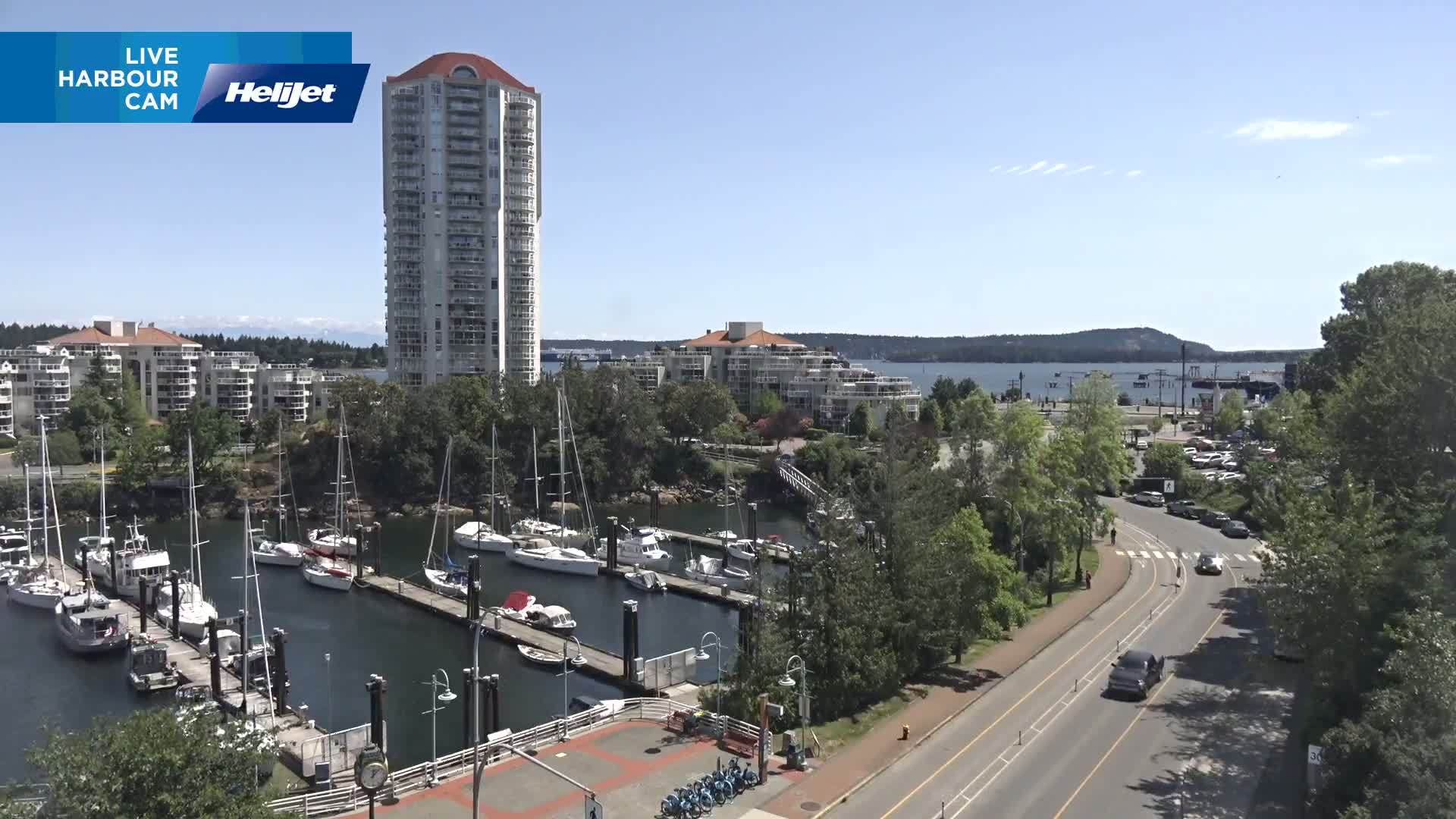 A sunny day reveals a marina filled with sailboats, a tall residential building, and a road with cars, all situated near a body of water with mountains in the distance.
