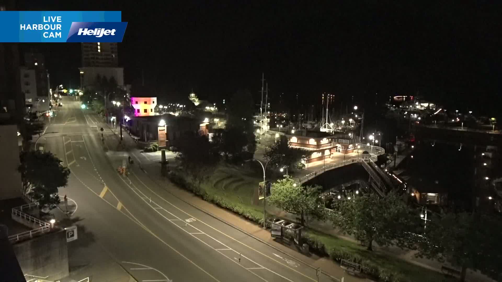 A nighttime, high-angle view shows a mostly empty harbor, a roadway, and several buildings with lights on, under clear skies.