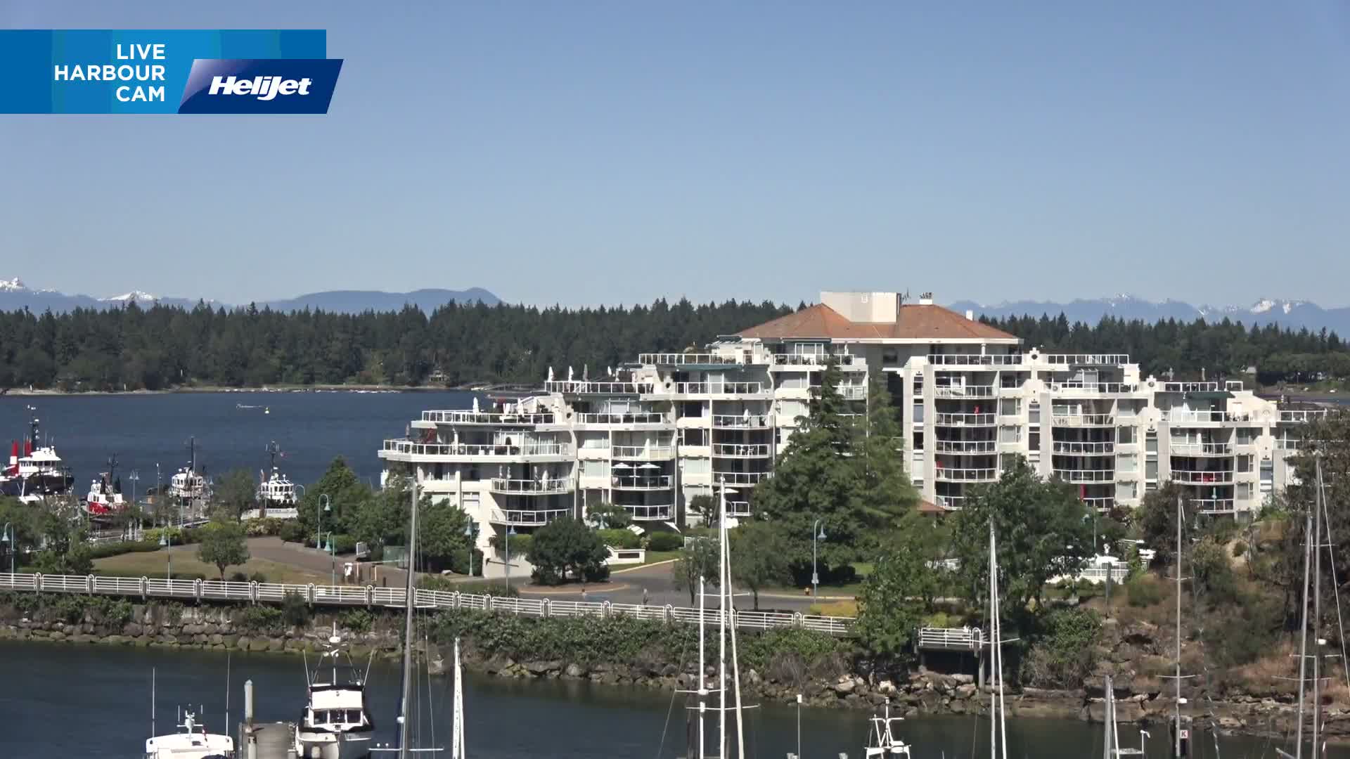 A large multi-story building situated on a waterfront, with several boats docked nearby, under a clear sunny sky and mountains in the background.