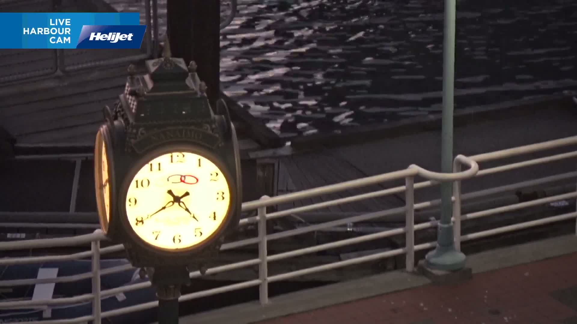 An illuminated clock mounted on a post stands beside a railing overlooking dark, choppy water at dusk.