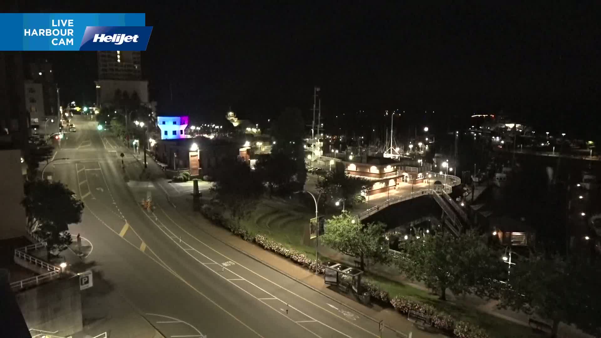 A nighttime view of a harbor with boats docked, a roadway with minimal traffic, and surrounding buildings illuminated under a clear, dark sky.