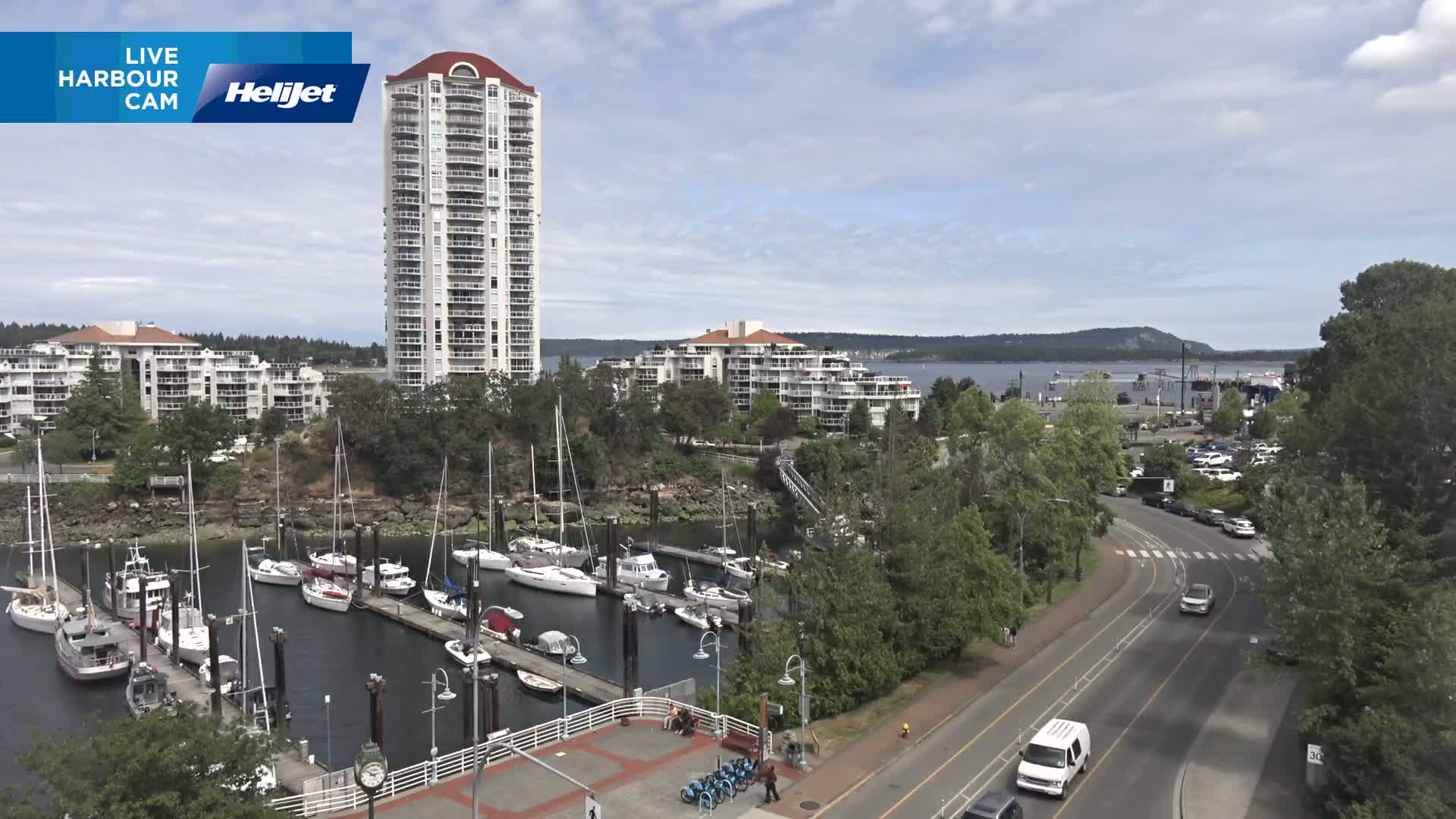 A marina filled with sailboats and motorboats sits beside a paved road and tall apartment buildings under a partly cloudy sky.