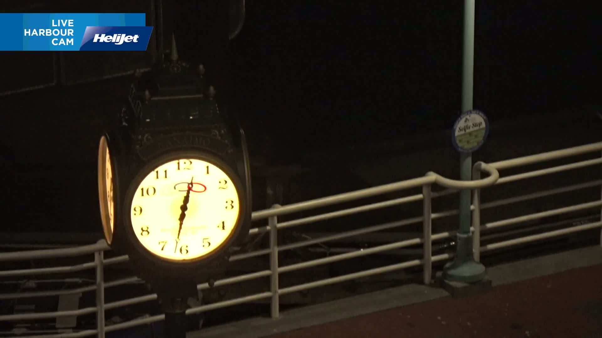 A nighttime view of a lighted clock mounted on a post next to a white railing.