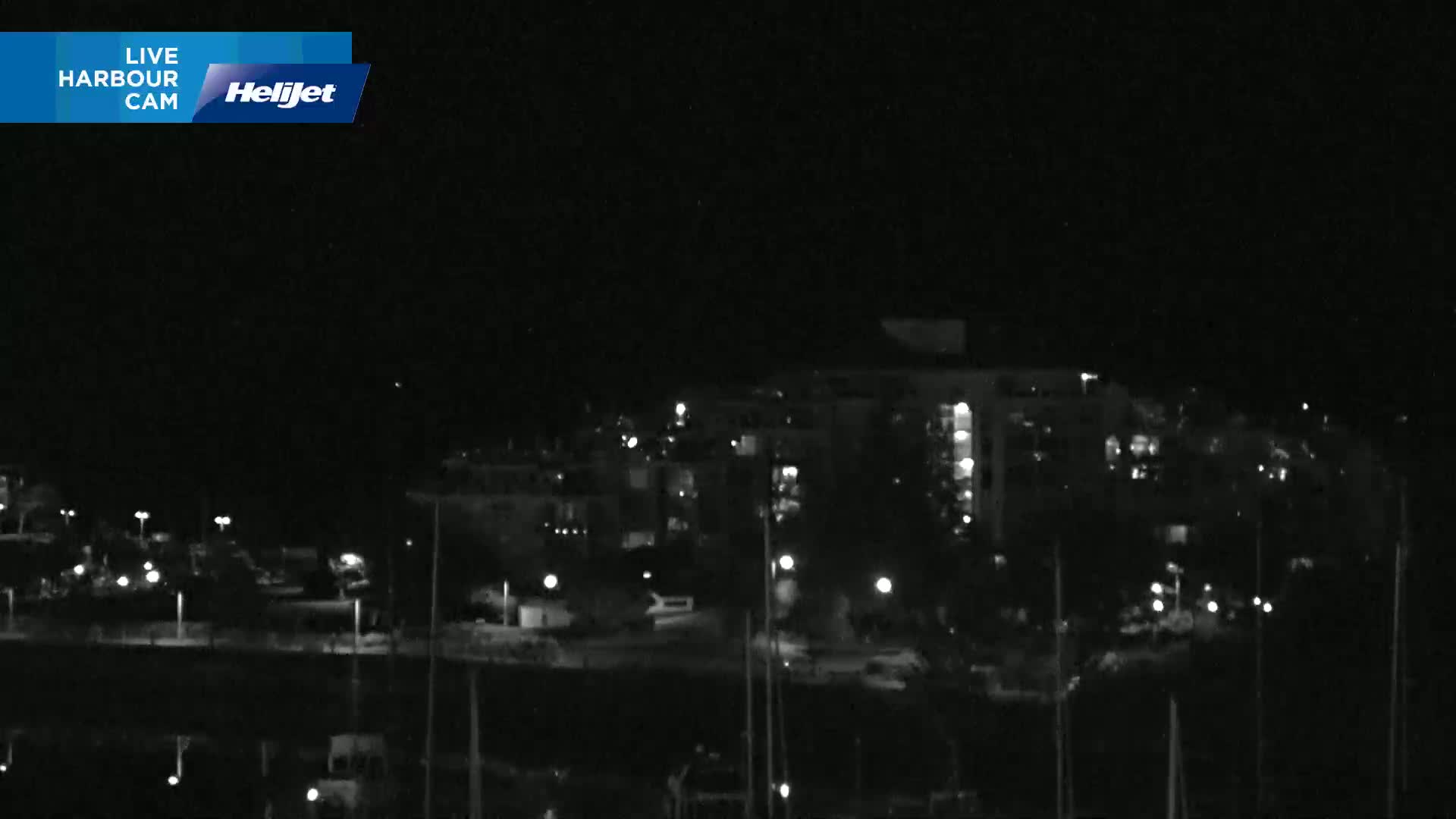 A nighttime view of a harbor area shows a multi-story building and dimly lit waterfront with boats moored.