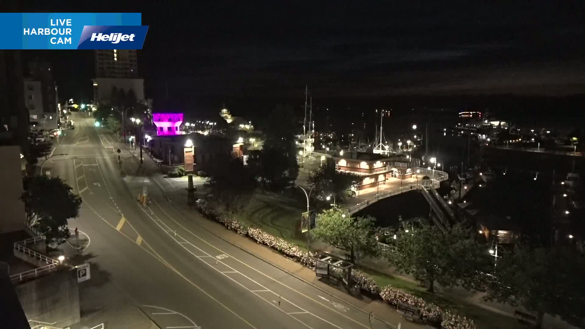 A nighttime aerial view shows a harbor with docked boats and a roadway with minimal traffic, under a clear, dark sky.