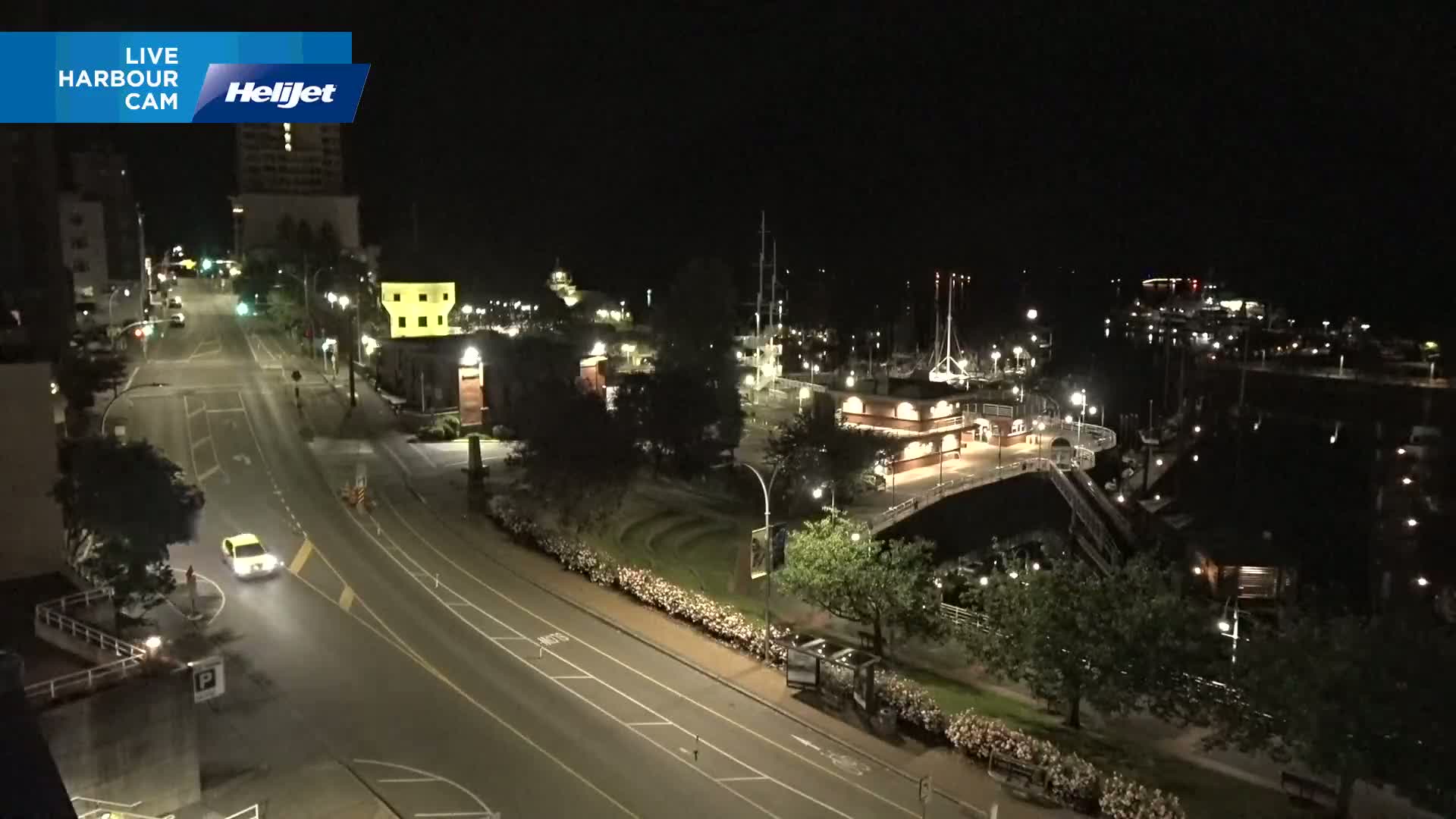 A nighttime, high-angle view shows a harbor with docked boats, a roadway with a single vehicle, and surrounding buildings and landscaping illuminated by streetlights.