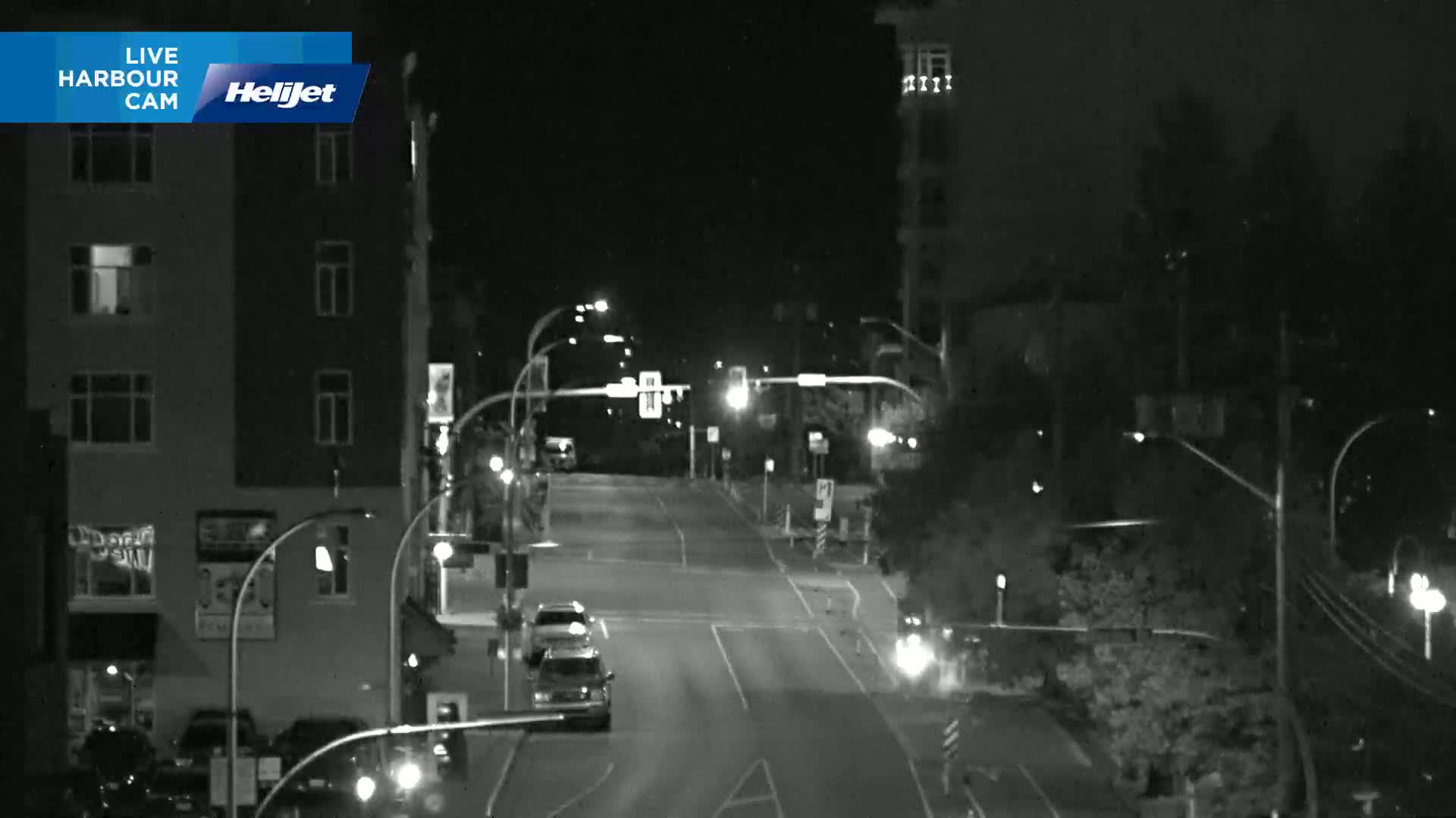 A nighttime, black-and-white view of a mostly empty intersection with several buildings and streetlights visible.