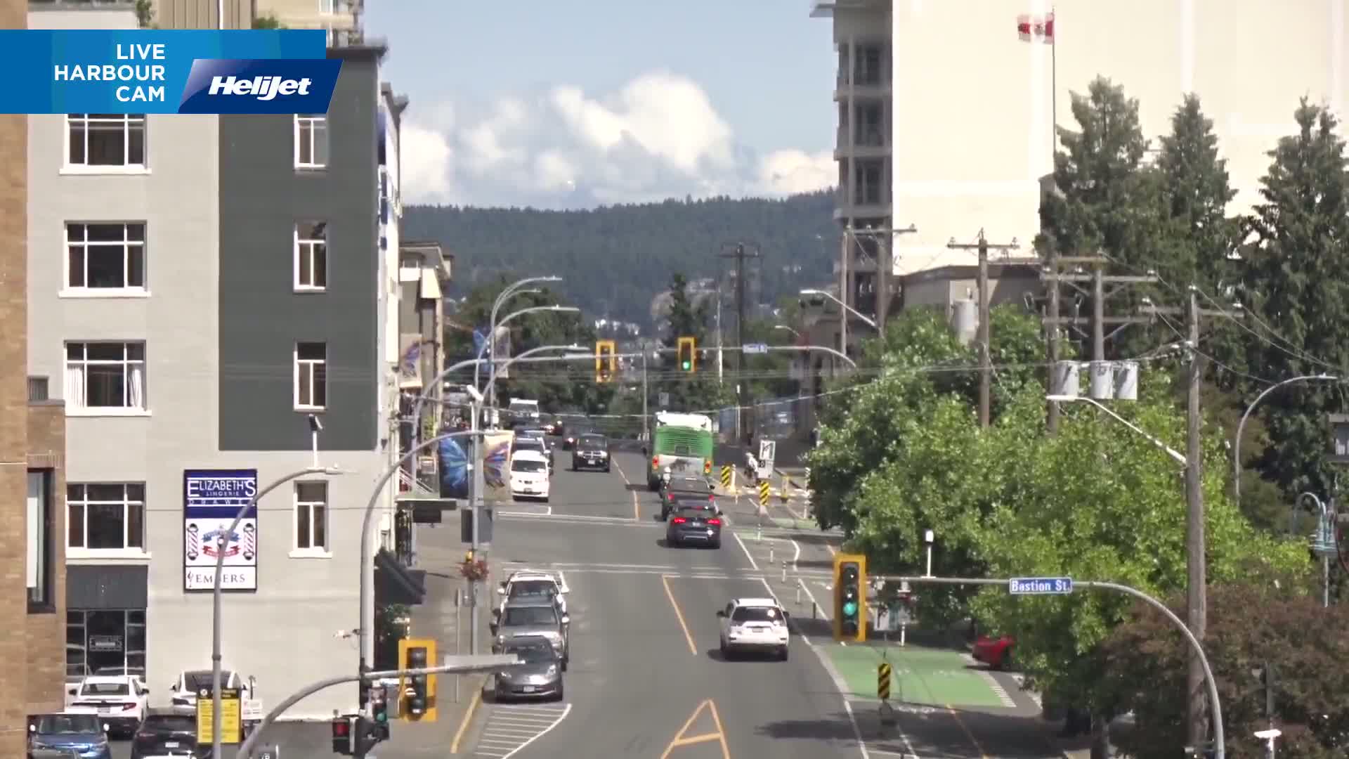 A sunny city street scene shows several cars driving on a road with a green bus, several buildings, and green trees lining the street, with mountains visible in the background.