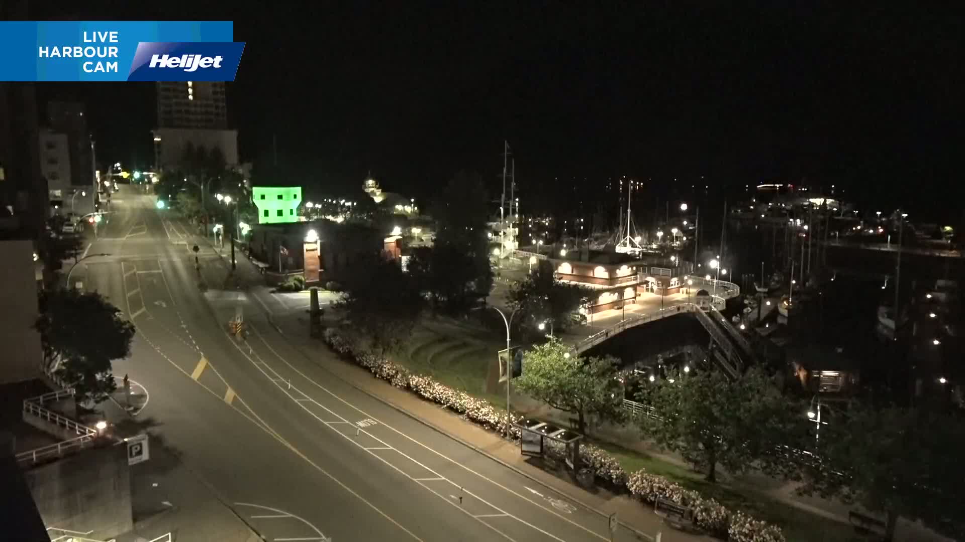 A nighttime view of a mostly empty harbor and roadway, with boats docked and streetlights illuminating the scene.
