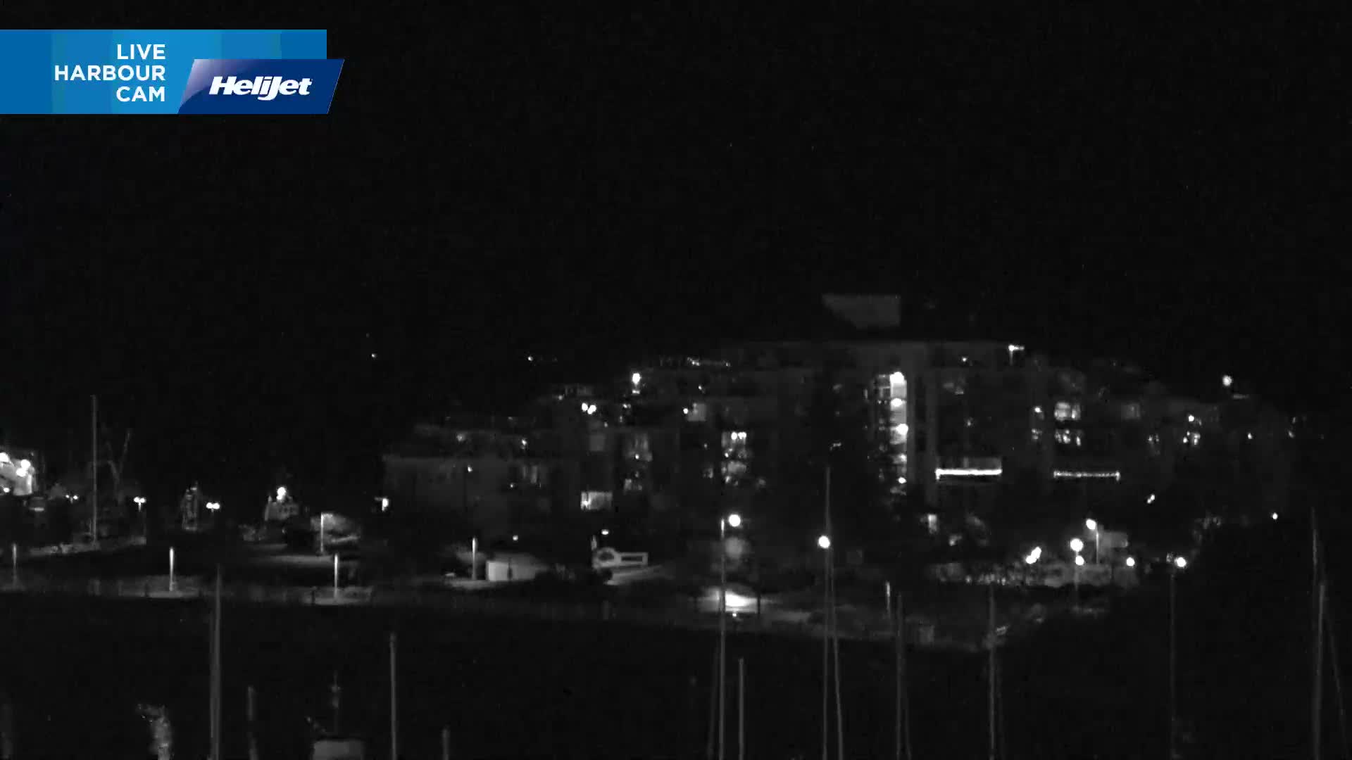A nighttime view of a harbor or marina with illuminated buildings and streetlights in the background, under clear skies.