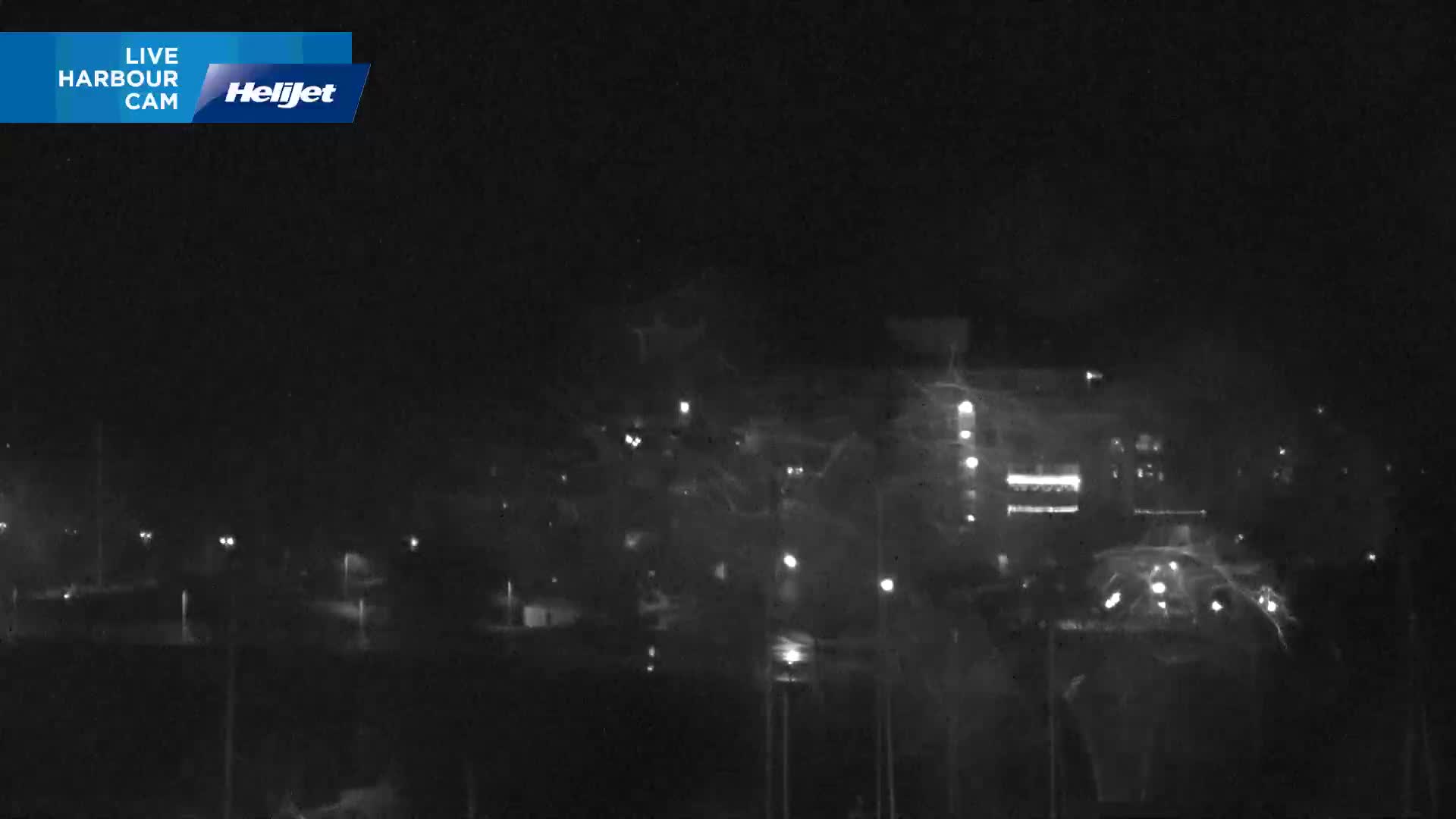 A blurry, dark nighttime view of a city or harbor is seen through a rain or moisture-covered camera lens, with numerous scattered lights indistinctly visible.