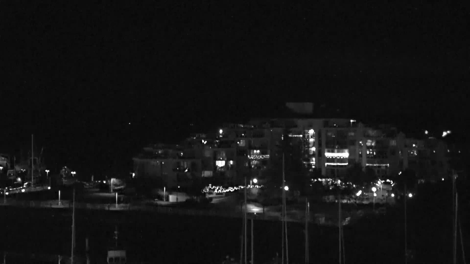 A nighttime scene reveals illuminated buildings and streetlights lining a waterfront, with numerous boat masts visible in the foreground under a clear, dark sky.