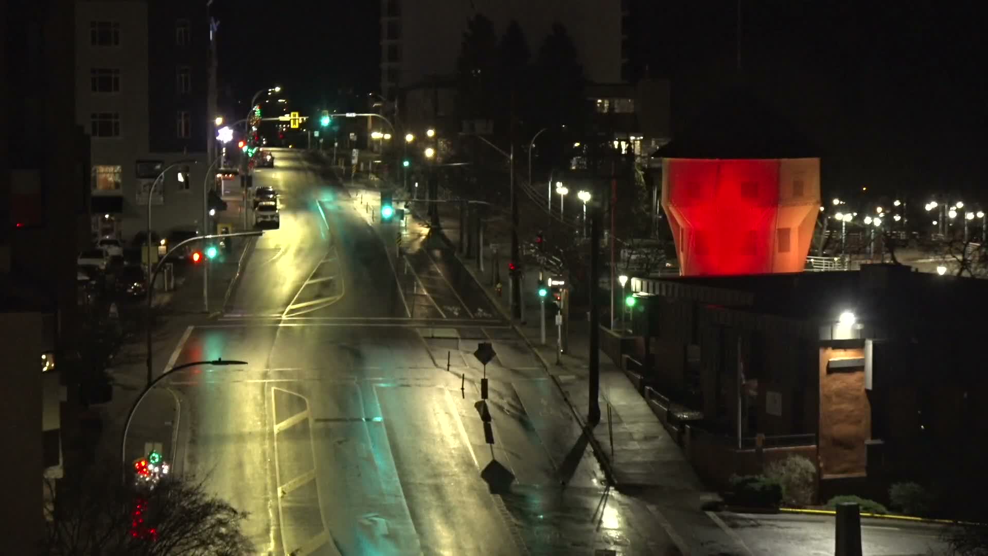 A nighttime scene reveals illuminated buildings and streetlights lining a waterfront, with numerous boat masts visible in the foreground under a clear, dark sky.
