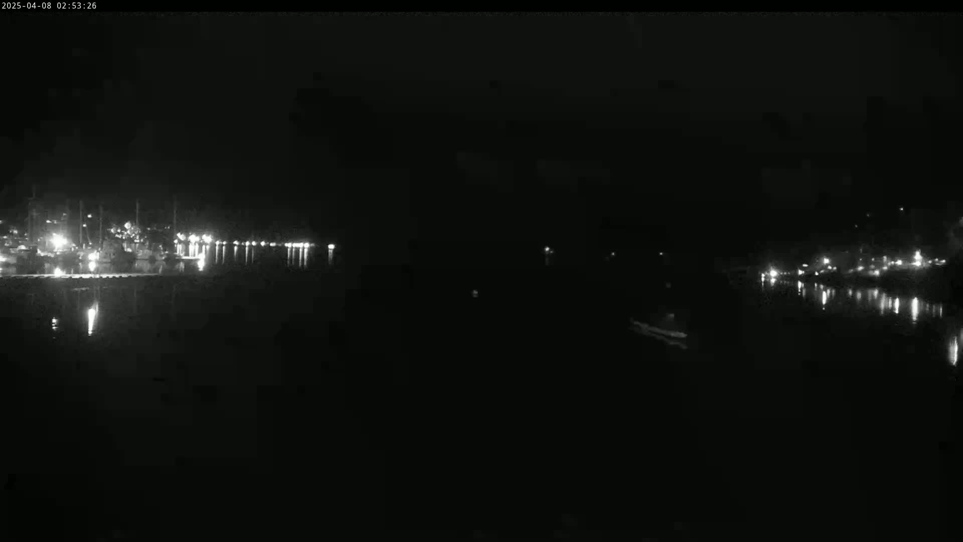 A nighttime, low-light image shows a dark body of water reflecting the lights of a distant shore with boats and buildings.