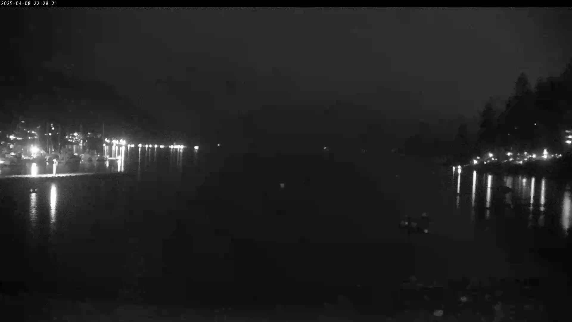 A nighttime, dark view of a body of water with lights reflecting on its surface from buildings and trees on either side under a hazy sky.