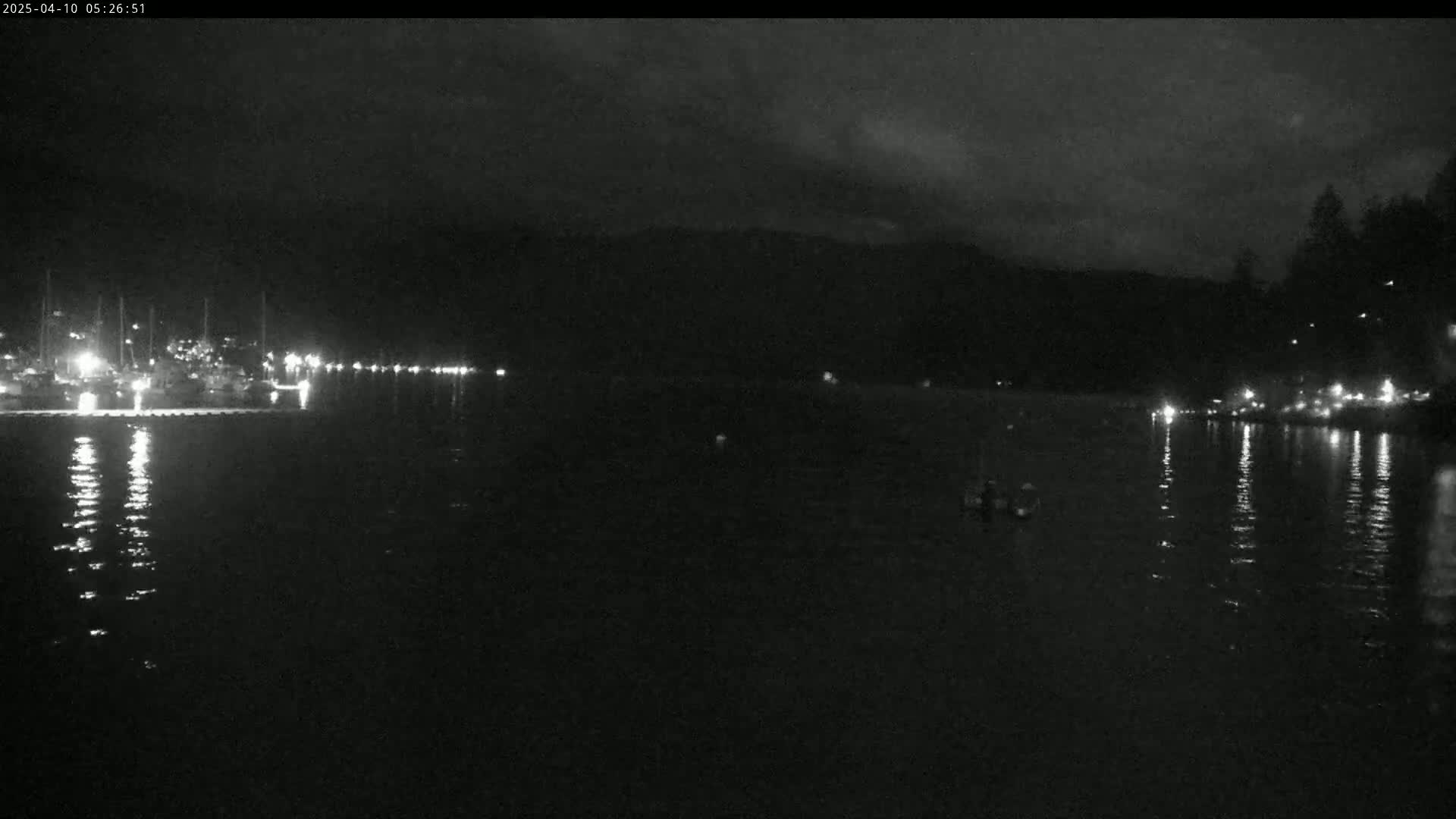 A dark, nighttime scene shows a body of water reflecting lights from a distant shoreline with boats and buildings.