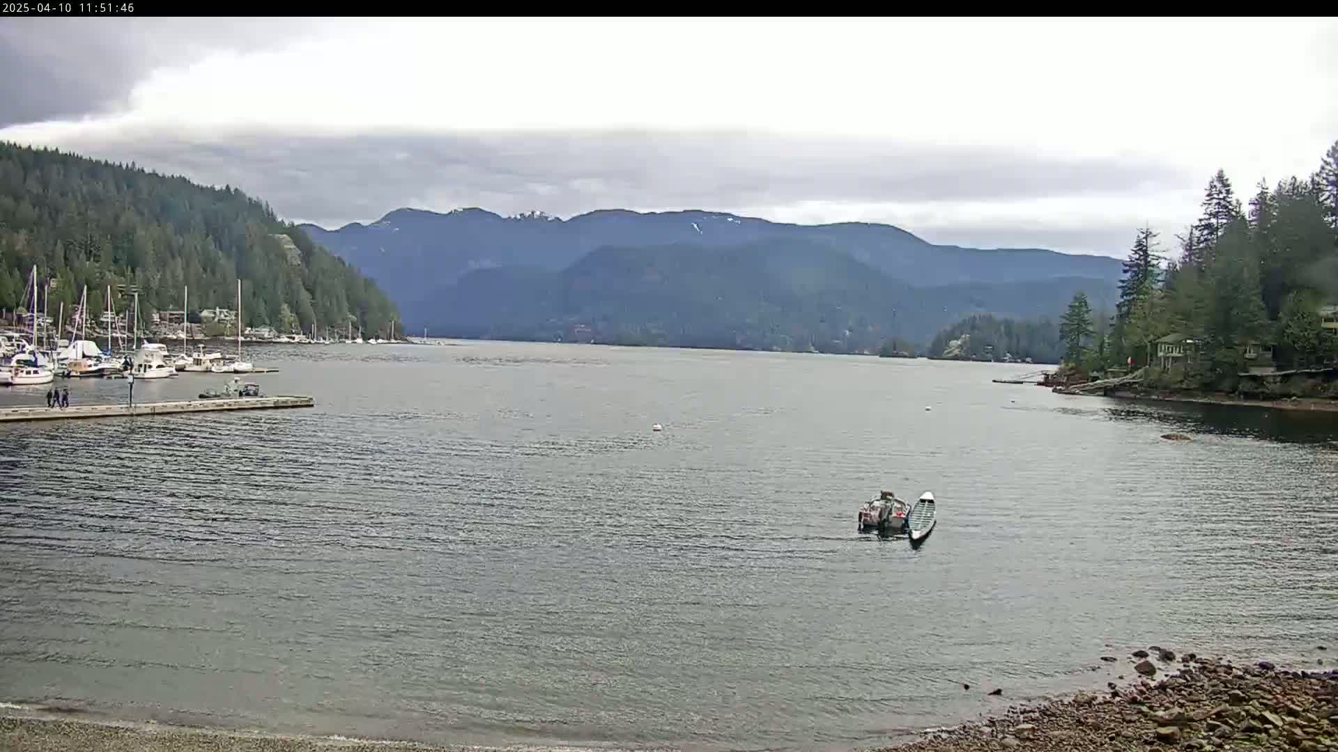 A calm bay with a few boats and a dock, surrounded by lush green mountains under an overcast sky.
