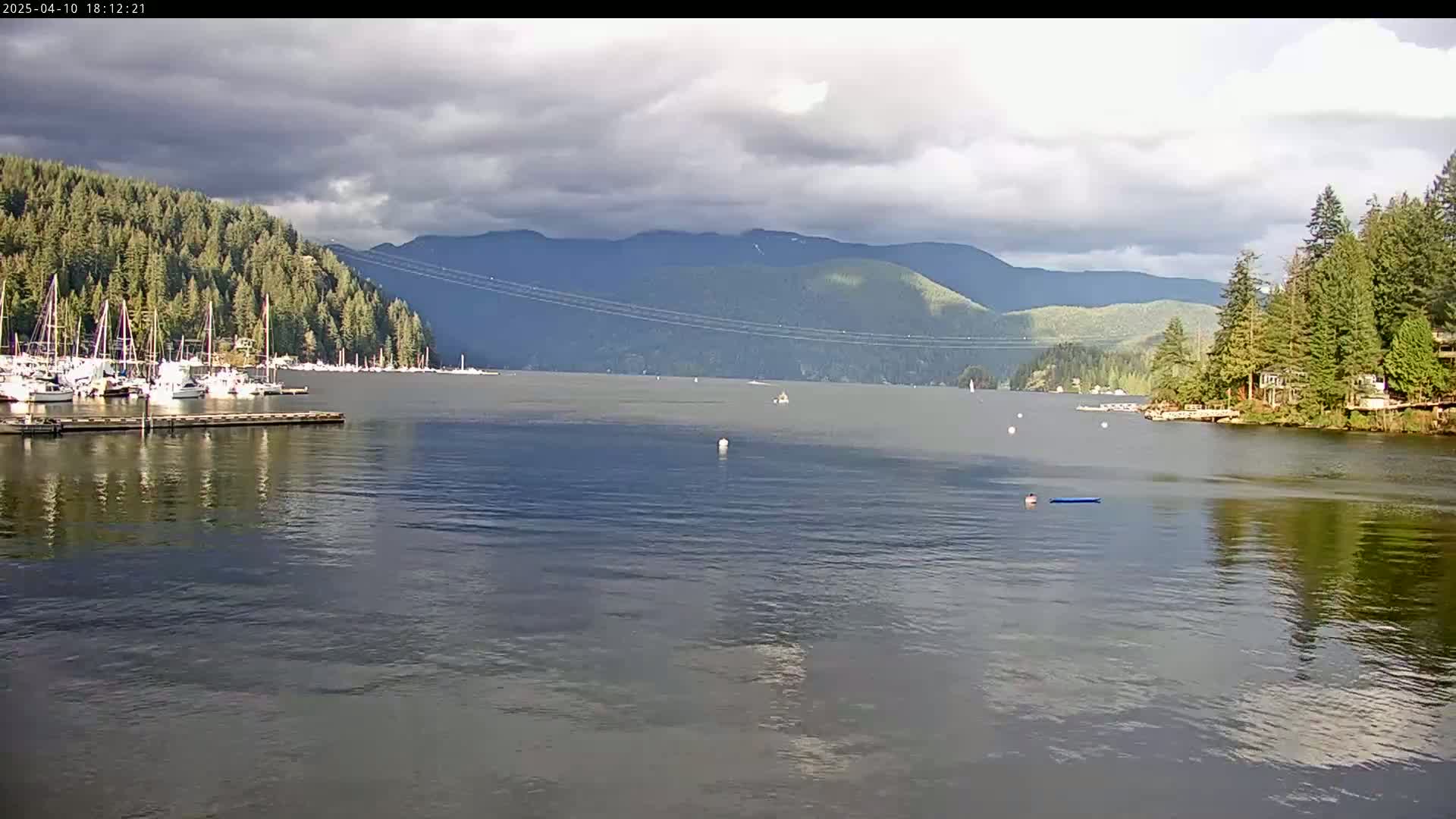 A calm body of water with numerous sailboats docked near a forested shoreline, mountains in the background under a partly cloudy sky.