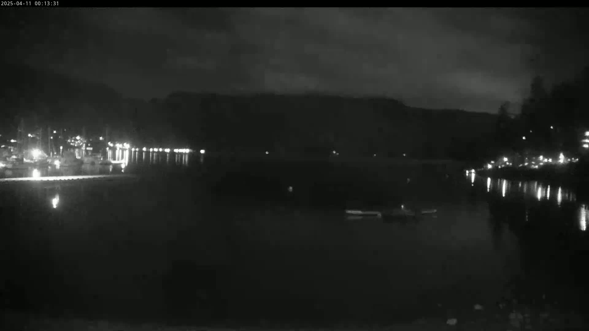 A nighttime, low-light view of a calm body of water reflecting lights from a shoreline with buildings and trees in the background under a mostly cloudy sky.
