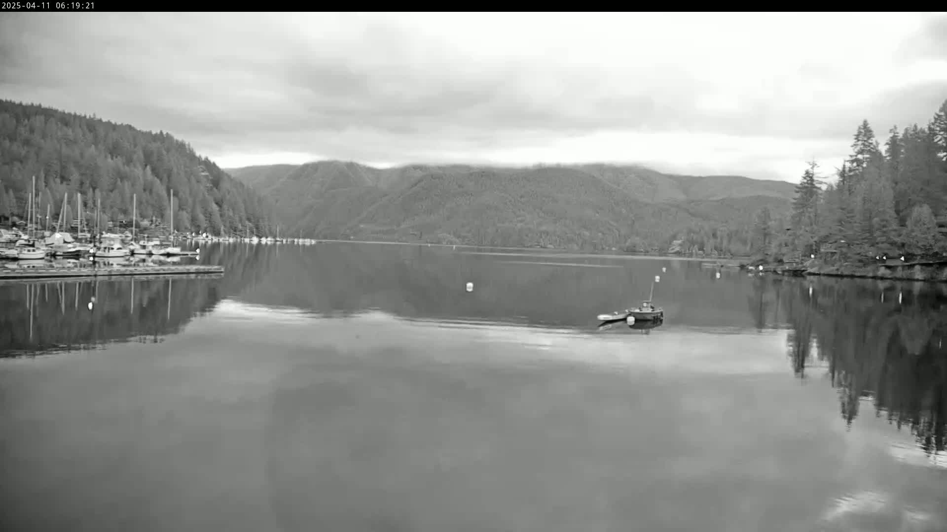 A calm, overcast day reveals a tranquil body of water reflecting forested hillsides, a small boat, and a marina filled with docked sailboats.