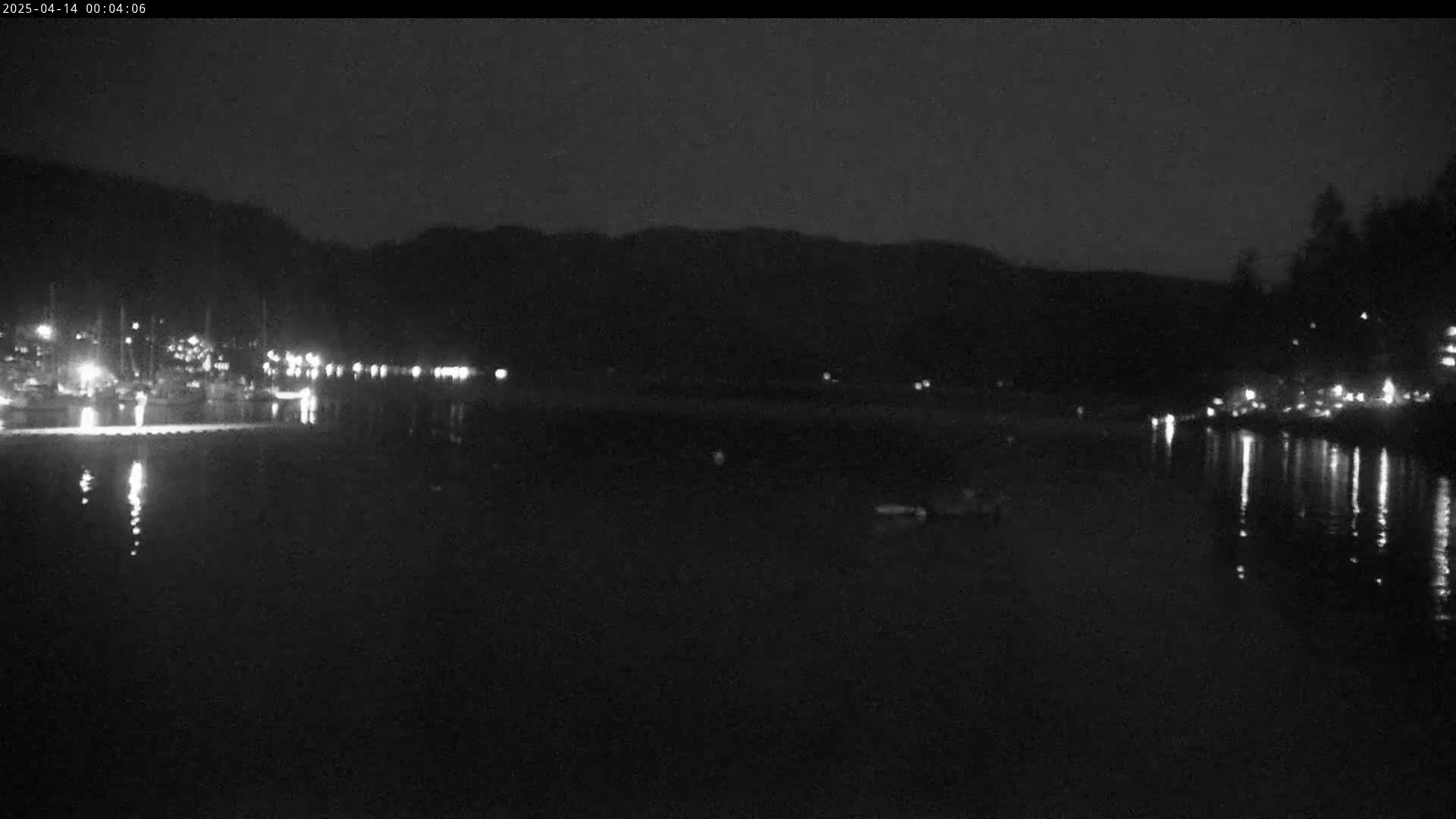 A nighttime, low-light image shows a calm body of water reflecting the lights from boats docked along a shoreline, with dark hills in the background.