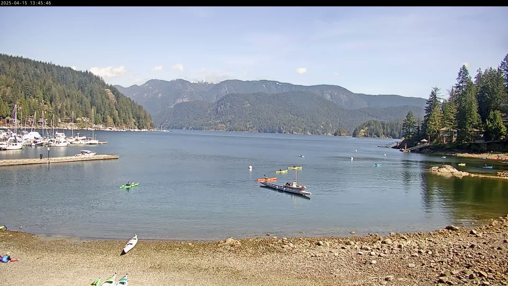 Kayakers paddle on a calm bay, surrounded by a rocky shore, a marina with numerous boats, and a mountainous backdrop under a sunny sky.