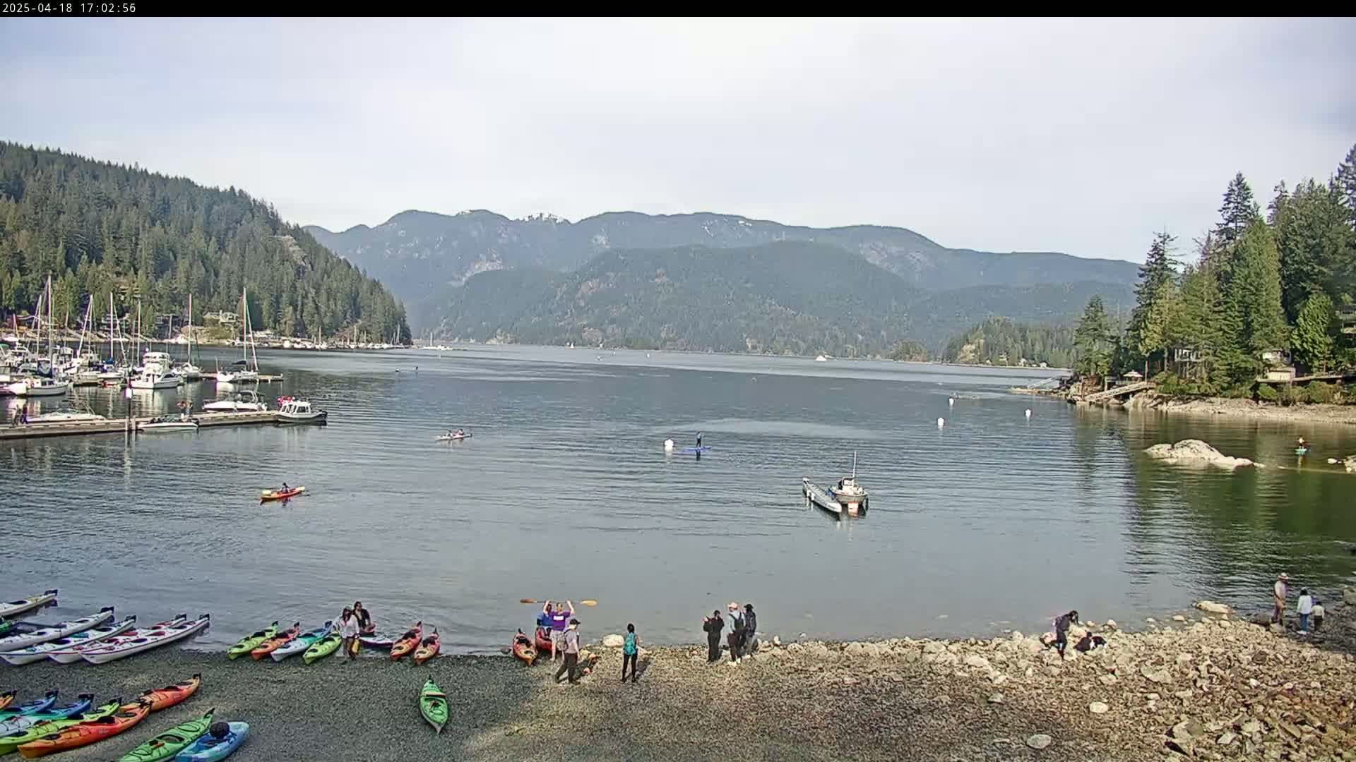 A calm bay surrounded by forested mountains under a mostly clear sky, with numerous kayaks and boats on the water and a few people gathered on a rocky shore.