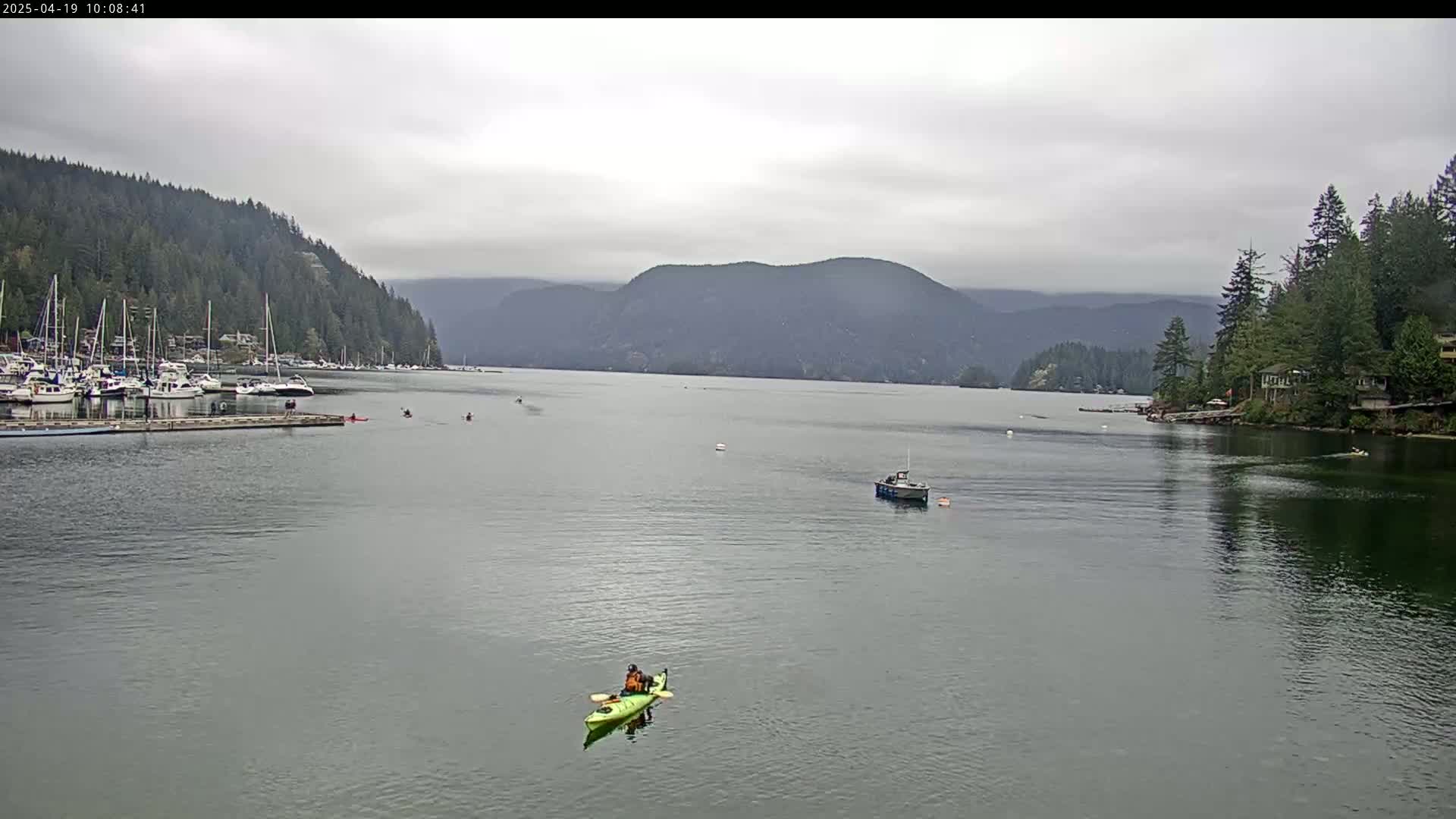 A calm, overcast day shows a bay with several boats moored at a dock, a few kayakers paddling, and a motorboat further out on the water, all surrounded by forested hills.