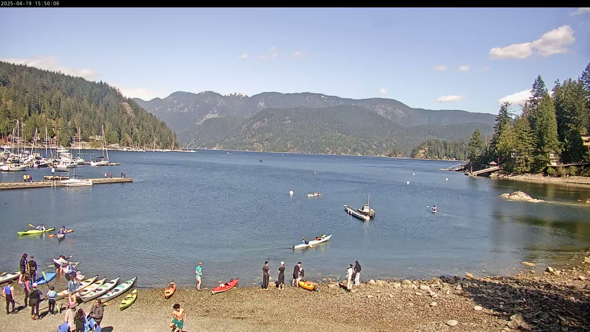A calm bay with a rocky shore, numerous kayaks and boats, and people on the beach, all set against a backdrop of forested mountains under a clear, sunny sky.