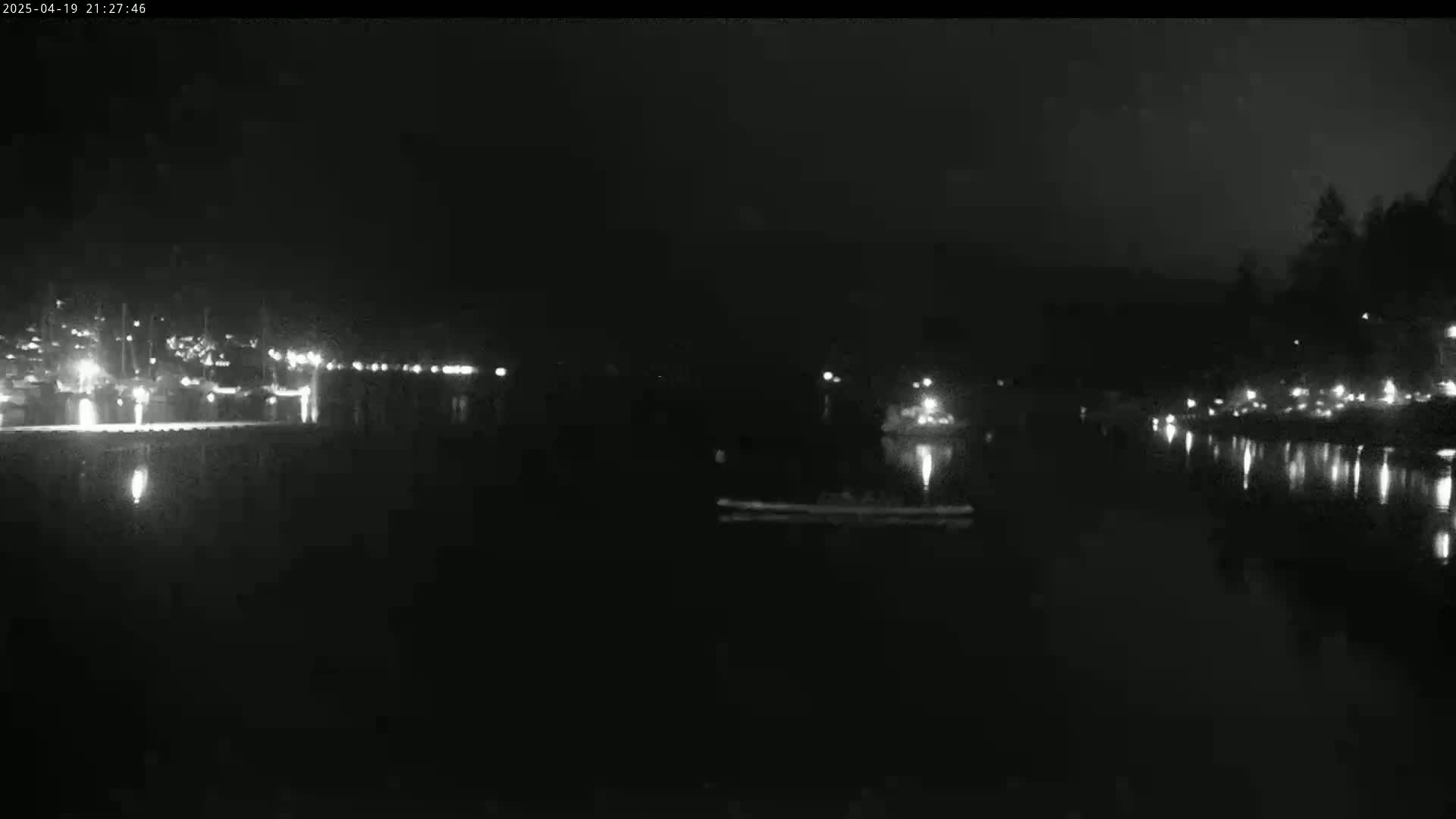 A nighttime view of a calm body of water with lights reflecting on its surface, showing boats and a shoreline with lights in the distance.