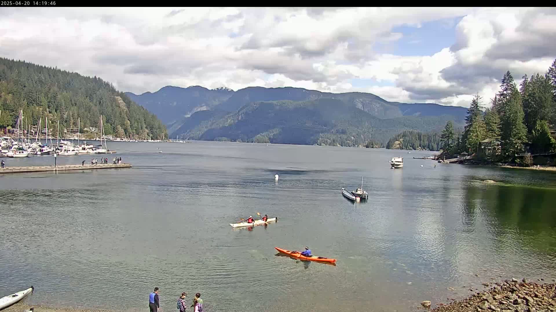 Kayakers paddle on calm water near a dock with boats and houses nestled amongst green mountains under a partly cloudy sky.