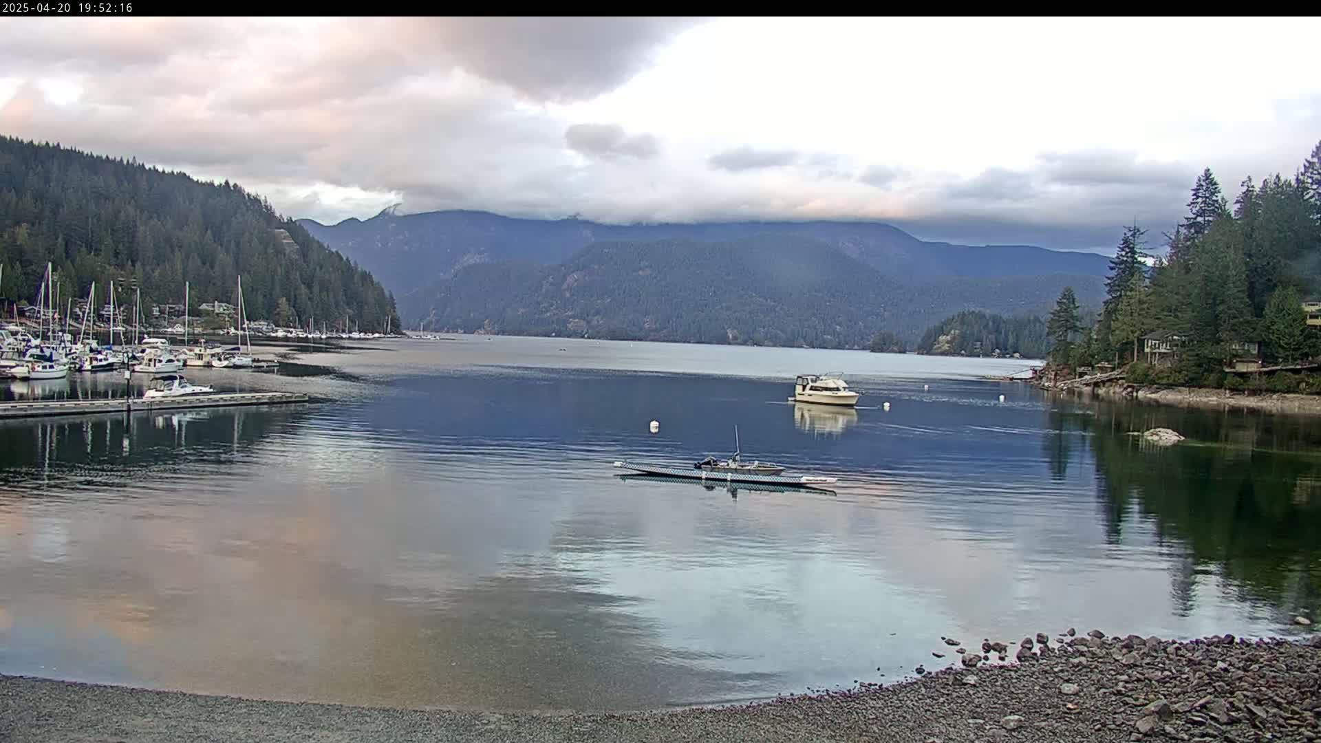 A calm bay with several boats at a marina is nestled between forested hills under a partly cloudy sky.