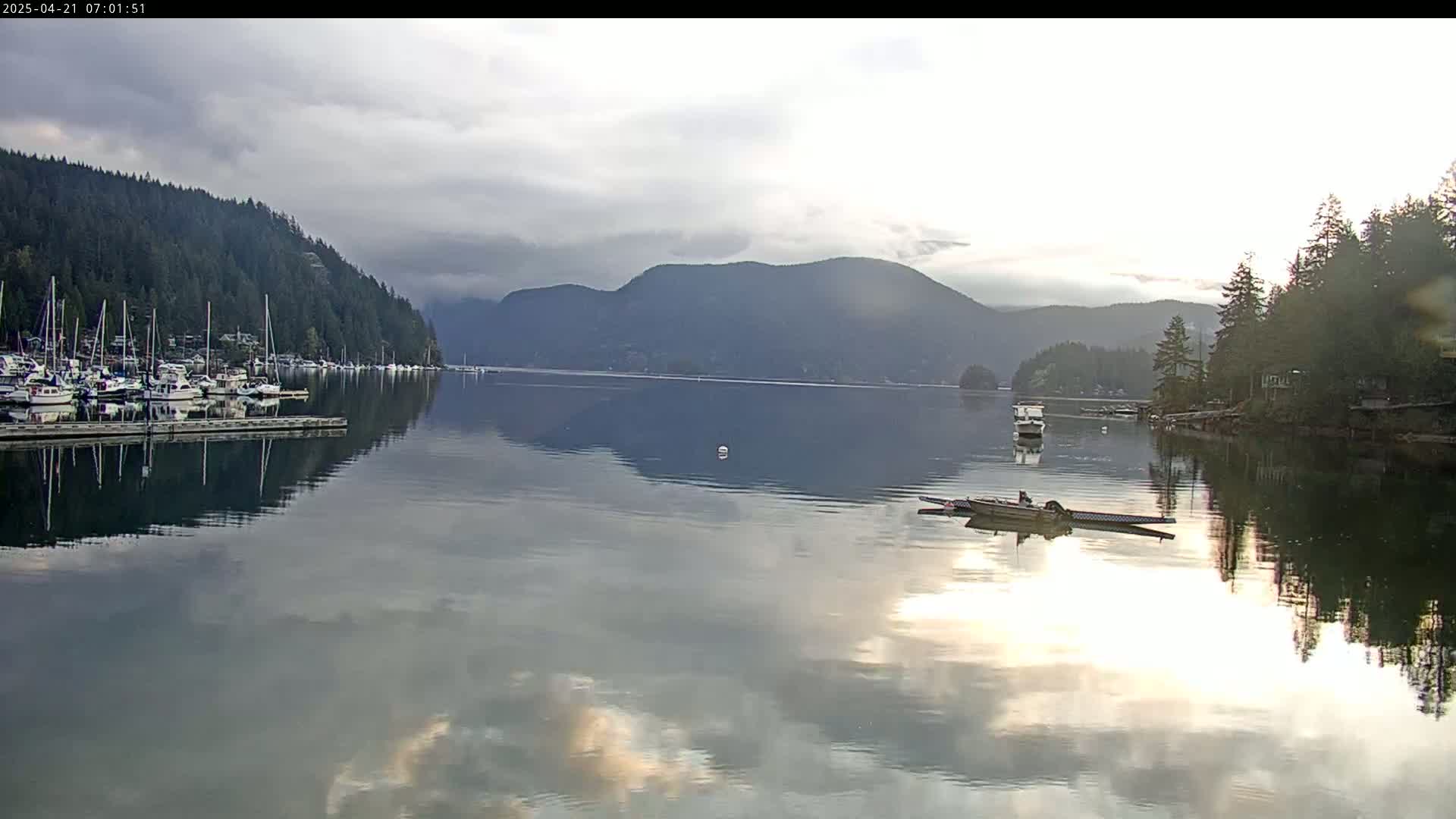 A calm bay with many boats docked at a marina, under a cloudy sky, reflects the surrounding mountains and trees.