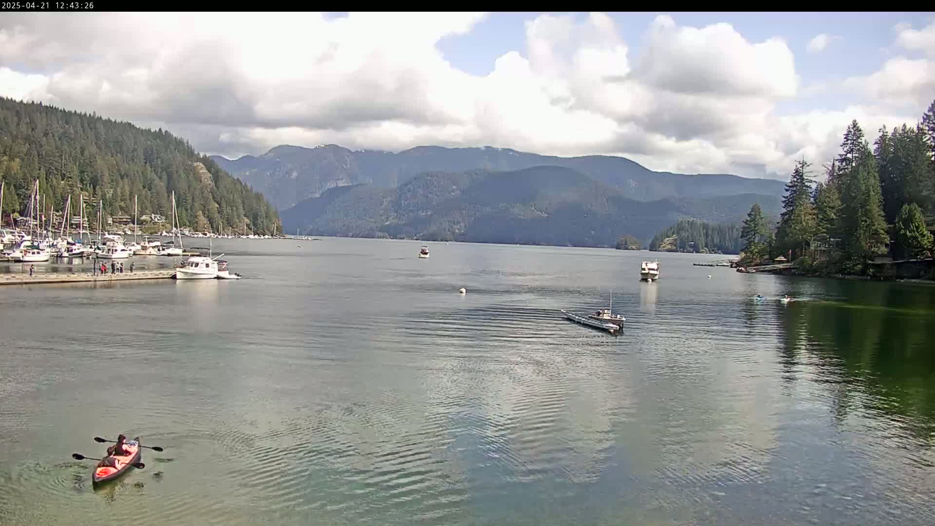 A calm bay with several boats and a kayak, partially cloudy skies, and mountains in the background.