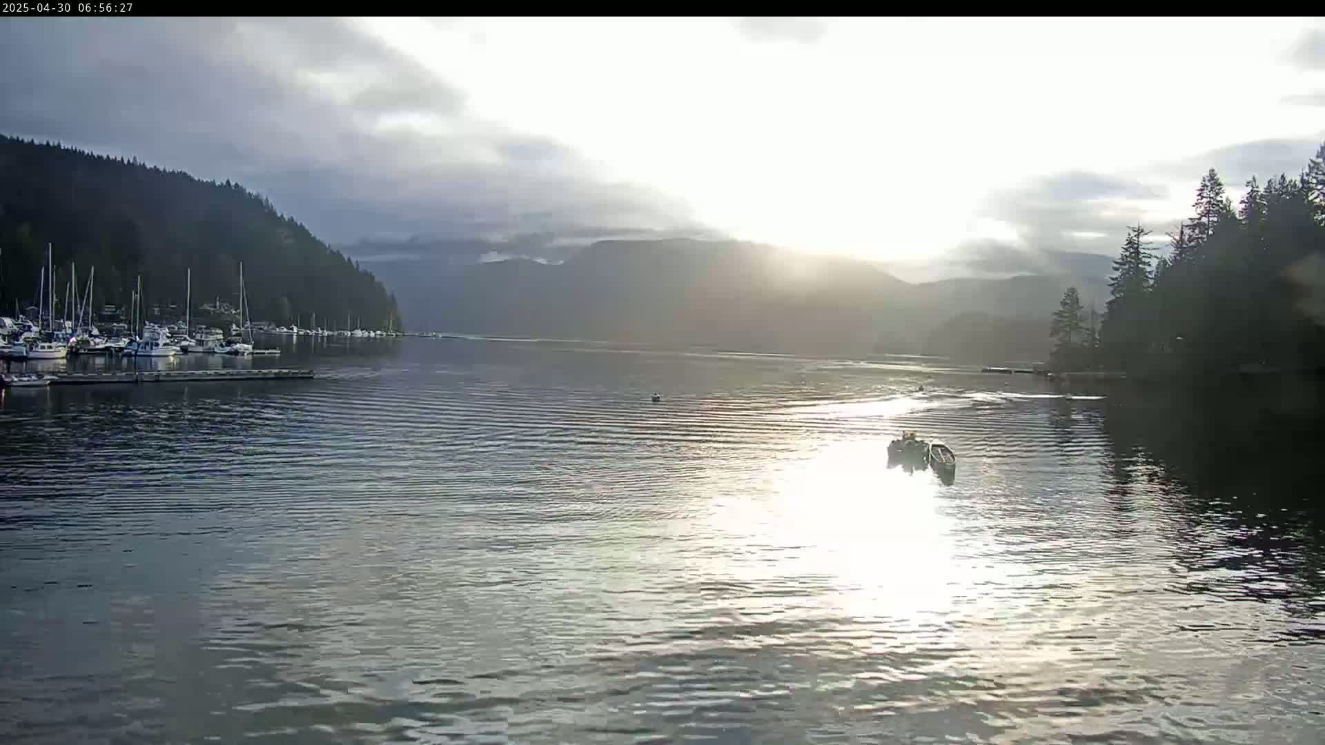 A calm body of water reflects the sunrise, with a marina full of boats to the left and a small boat with people in it in the center, all framed by forested hills.