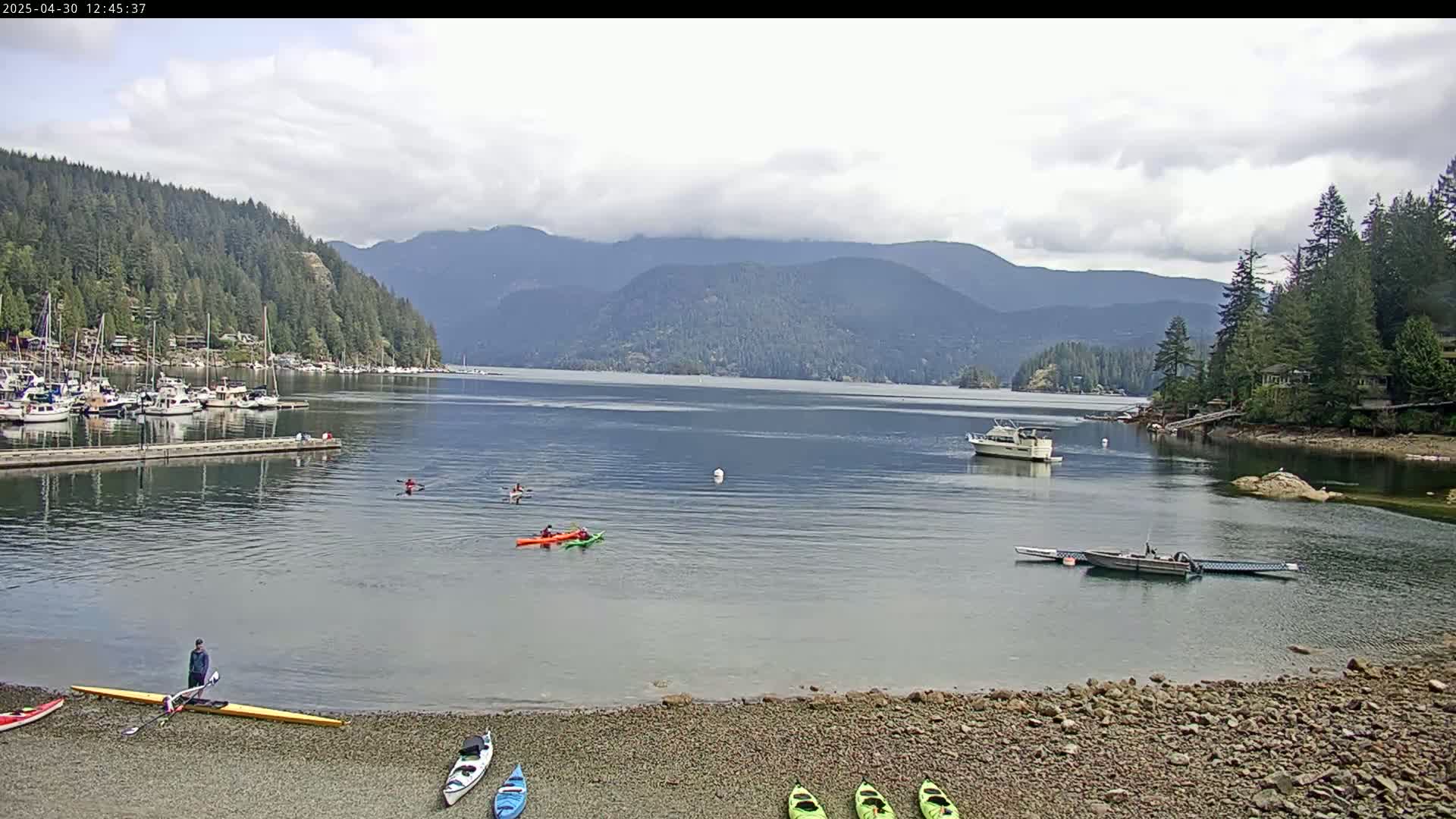 A calm bay with several kayaks and boats, a rocky shoreline, and forested mountains under a partly cloudy sky.