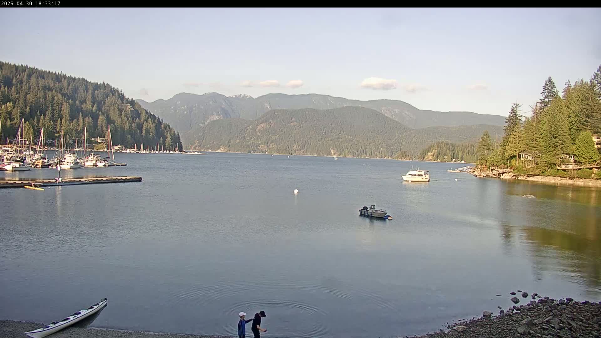 A calm bay, under a mostly clear sky, is surrounded by forested hills, with several boats moored in a marina and a few other boats on the water, and two people standing by the shore near a kayak.