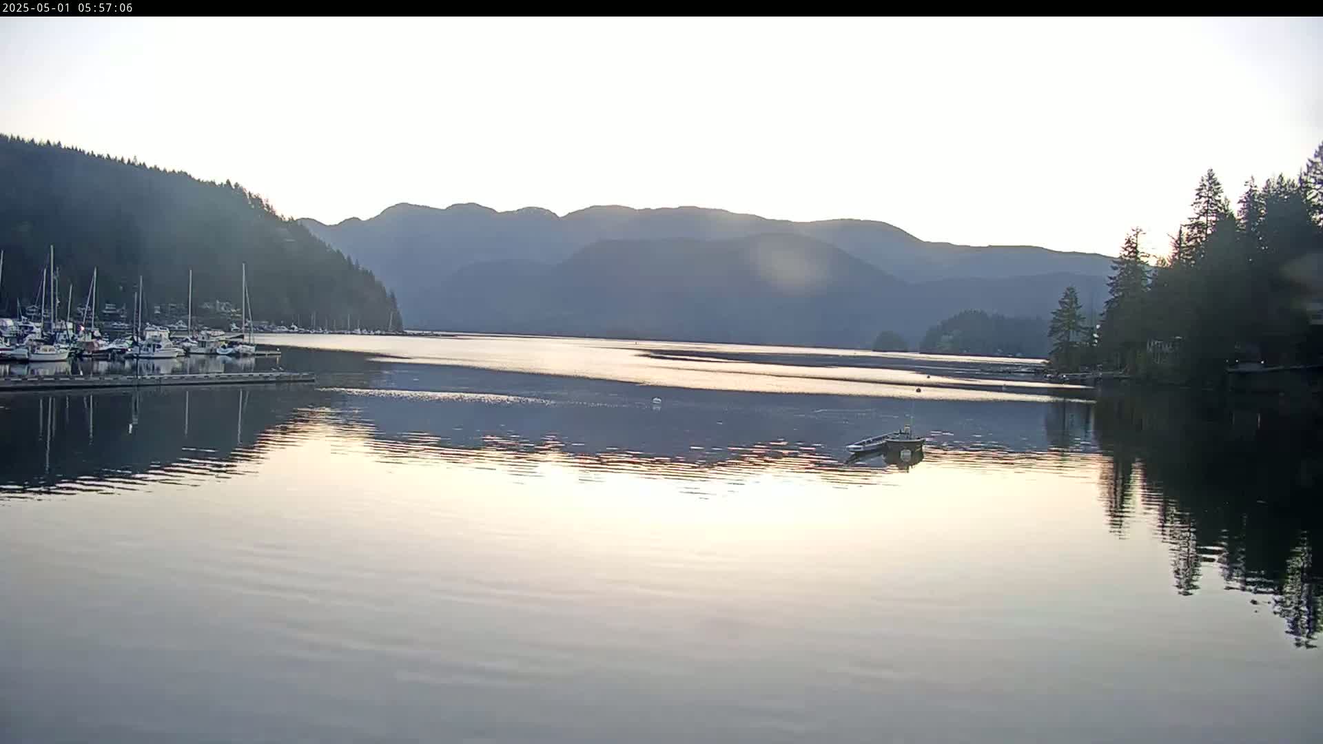 A calm body of water reflects a sunrise sky and mountains, with a marina full of boats to the left and forested shoreline to the right.