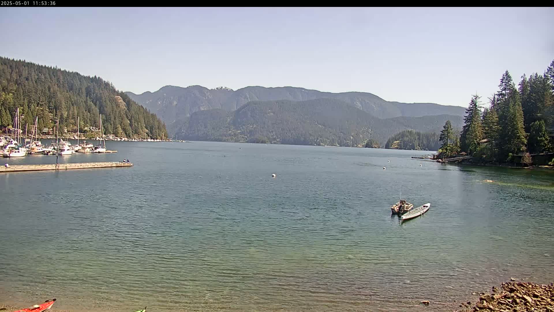 A calm, sunny day shows a bay with boats docked on one side and a few kayaks and a small motorboat on the water, all surrounded by forested mountains.