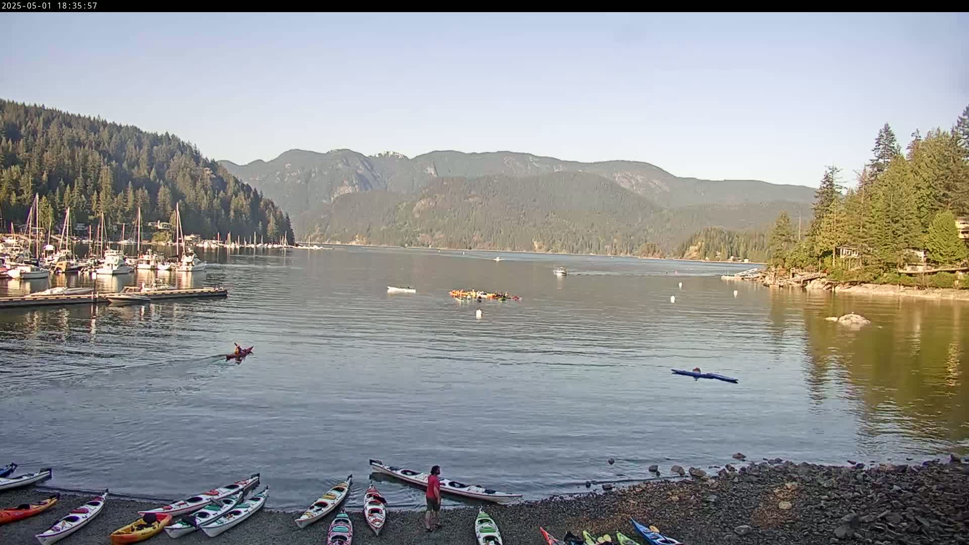 A calm bay with numerous boats docked, kayakers on the water, and kayaks lined up on a pebbled shore, all nestled against a backdrop of forested mountains under a clear sky.